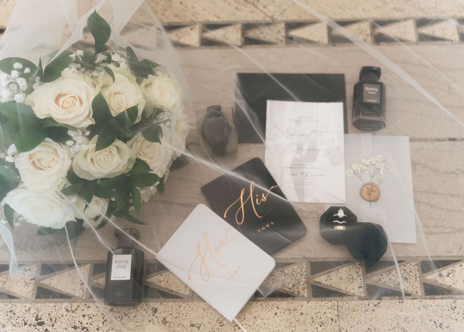 Elegant wedding flat lay with white roses, perfume, invitation cards, and ring on a marble surface.