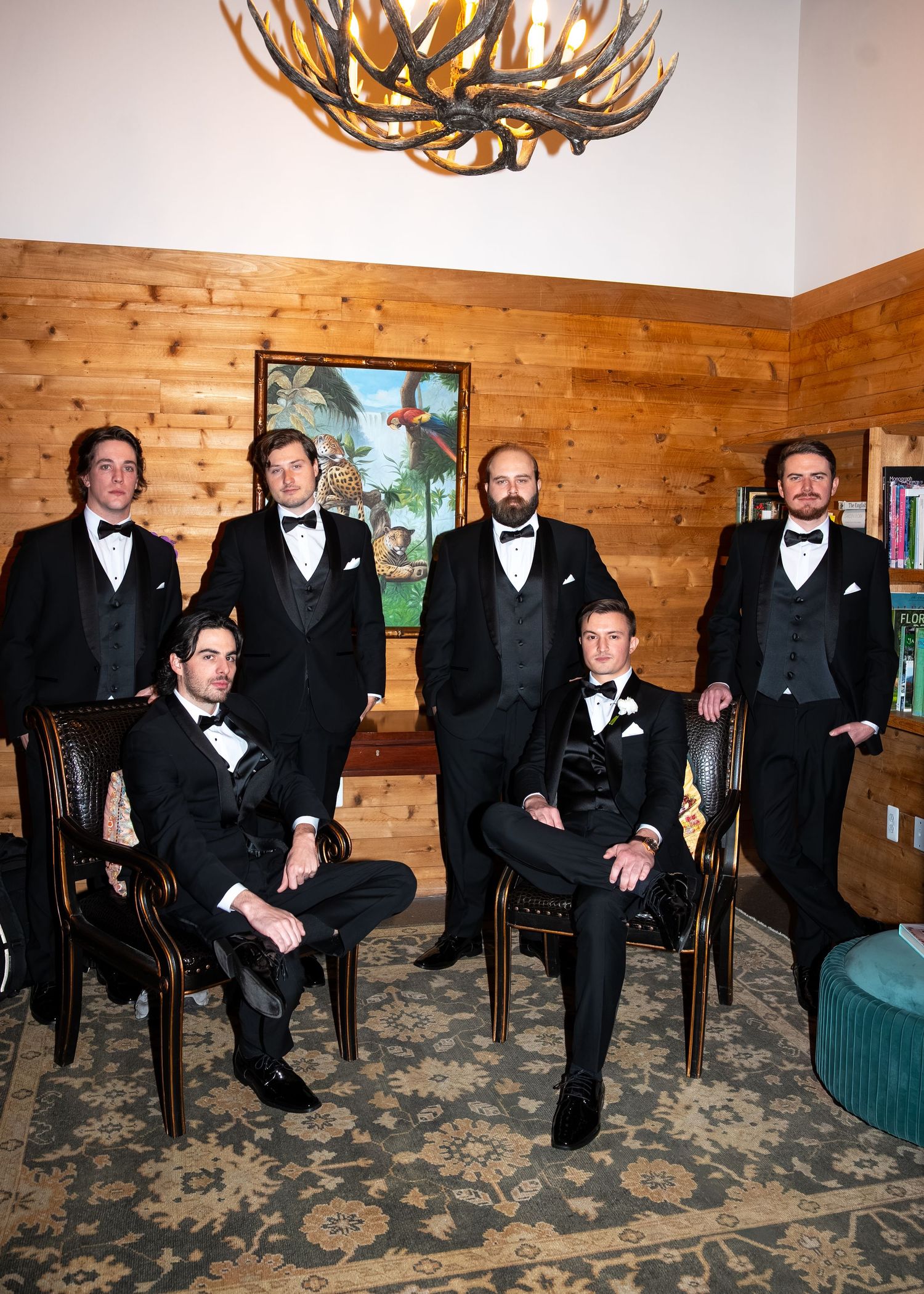 Six groomsmen in black tuxedos pose formally in a rustic wood-paneled room with antler chandelier.