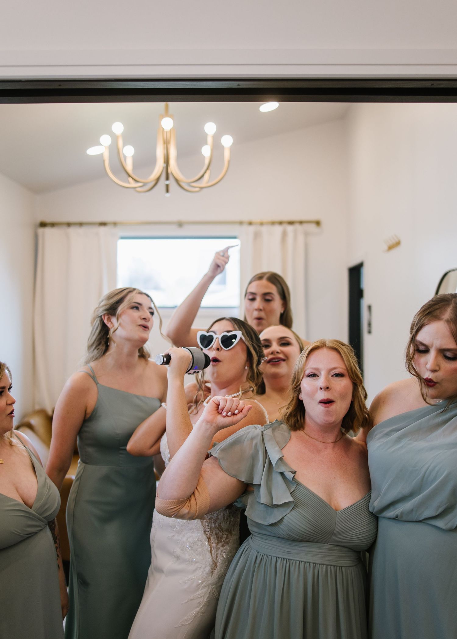 A group of friends in sage green dresses celebrate joyfully during a formal event under a chandelier.