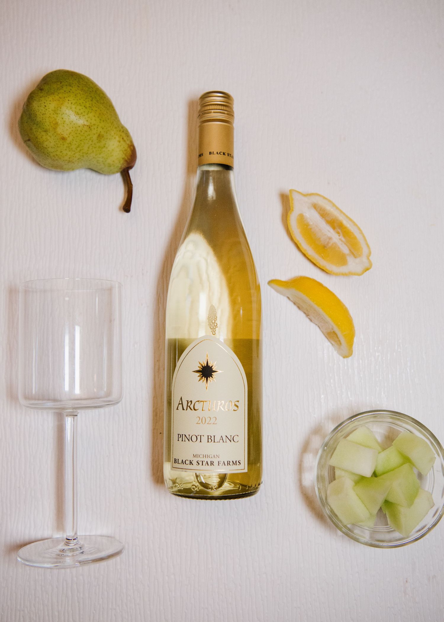 White wine bottle with pear, lemon wedges, wine glass and a cocktail glass filled with ice on white surface.
