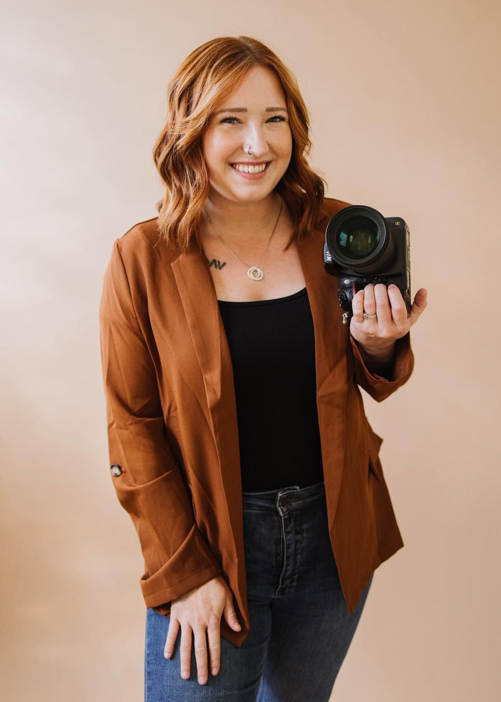 Smiling woman with red hair holding a camera, wearing a brown blazer and black top, posing against a beige background.