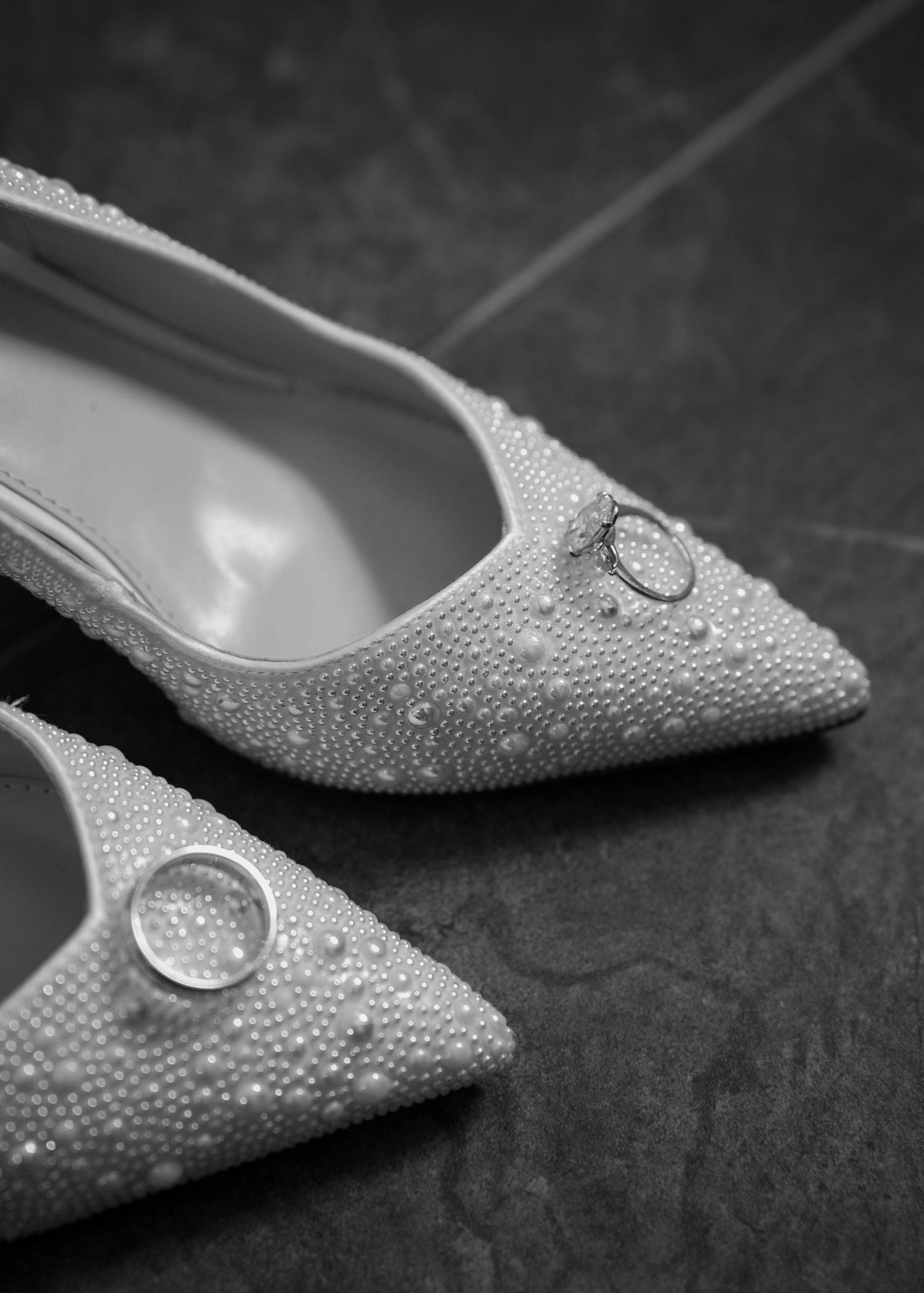 Black and white photo of pearl-encrusted bridal heels with wedding rings resting on the pointed toes.
