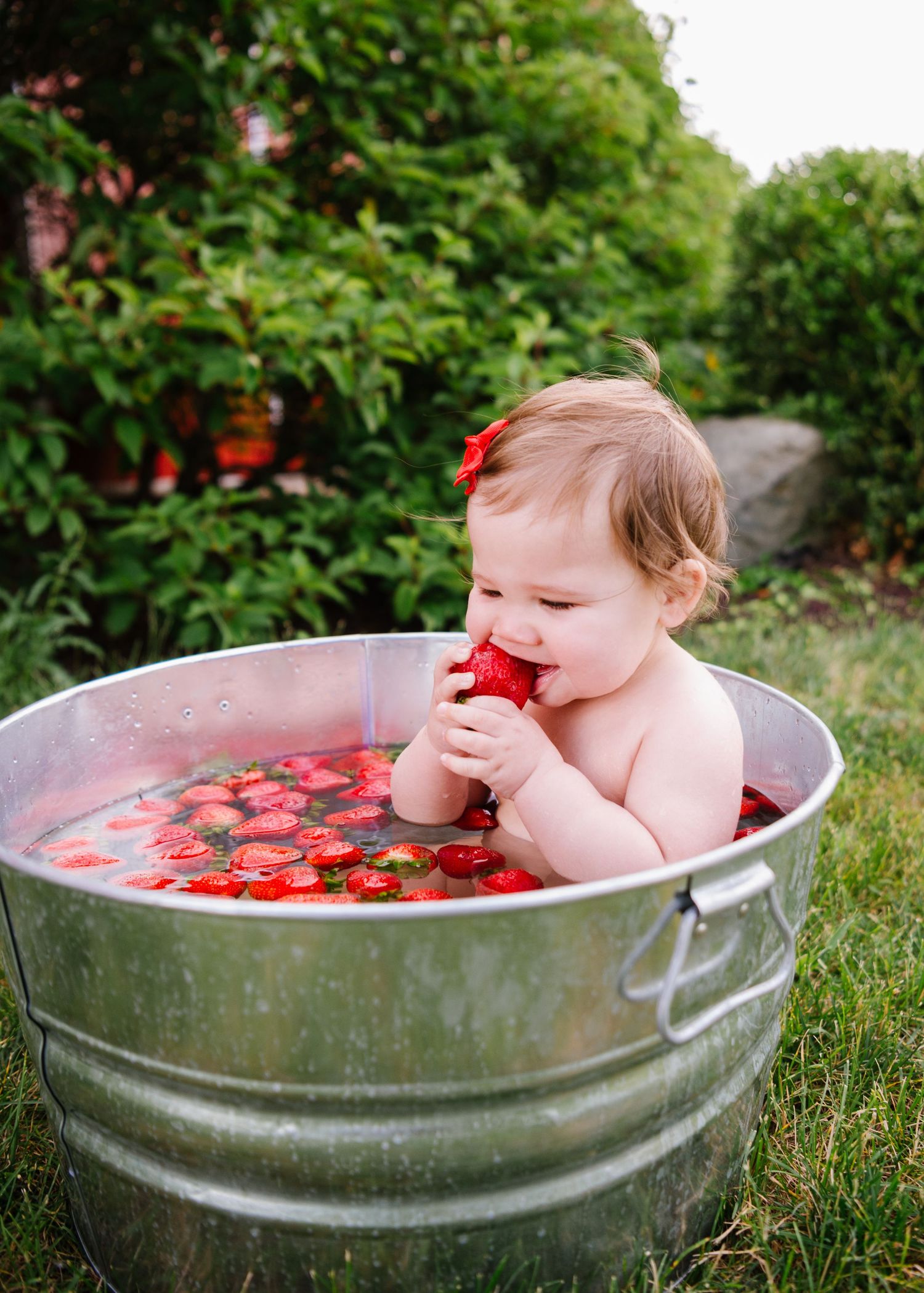 Small child sits in metal tub filled with floating strawberries and red-tinted water in outdoor garden setting.