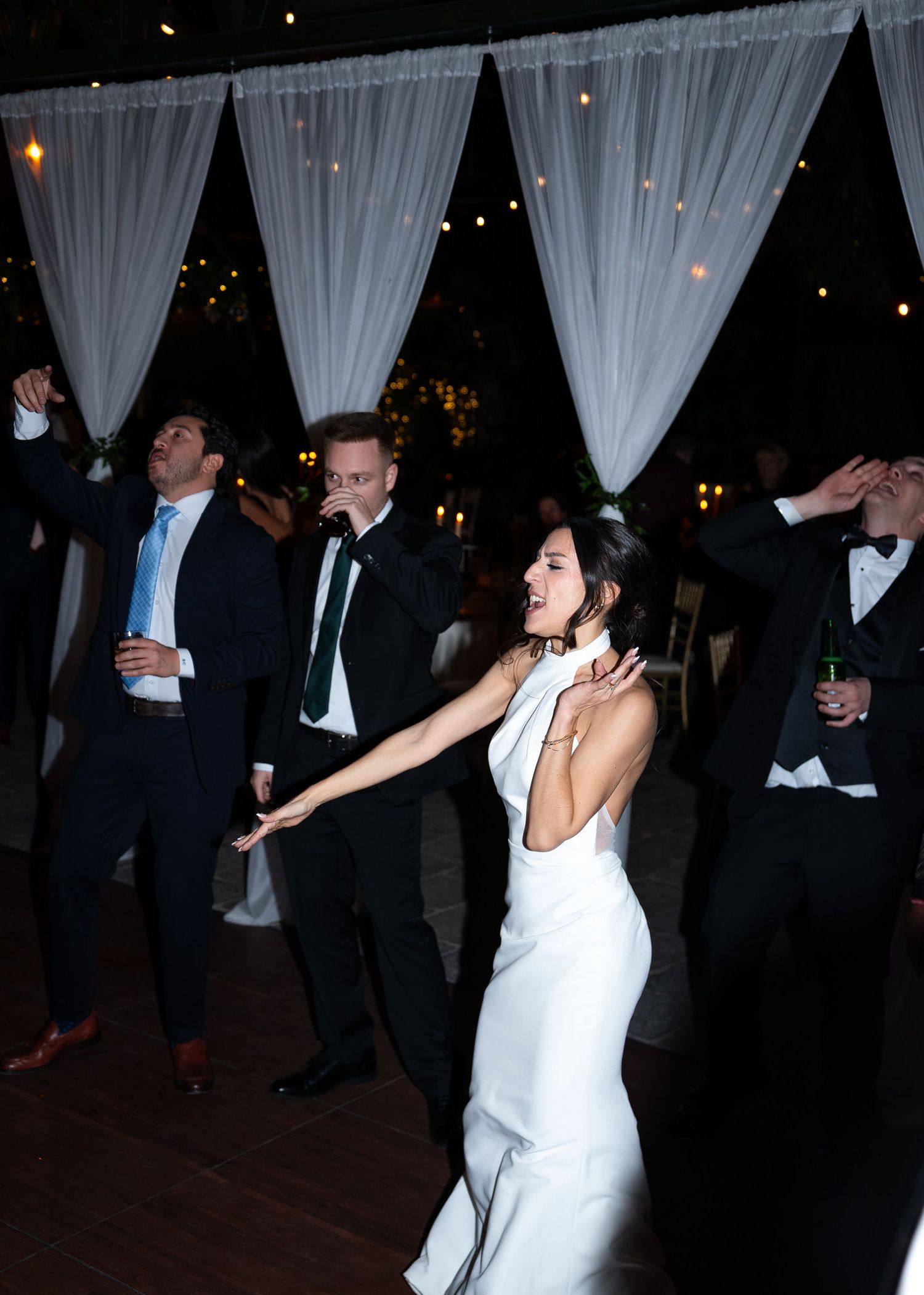 Bride in white gown dancing joyfully with guests in formal attire at an elegant outdoor wedding reception at night.