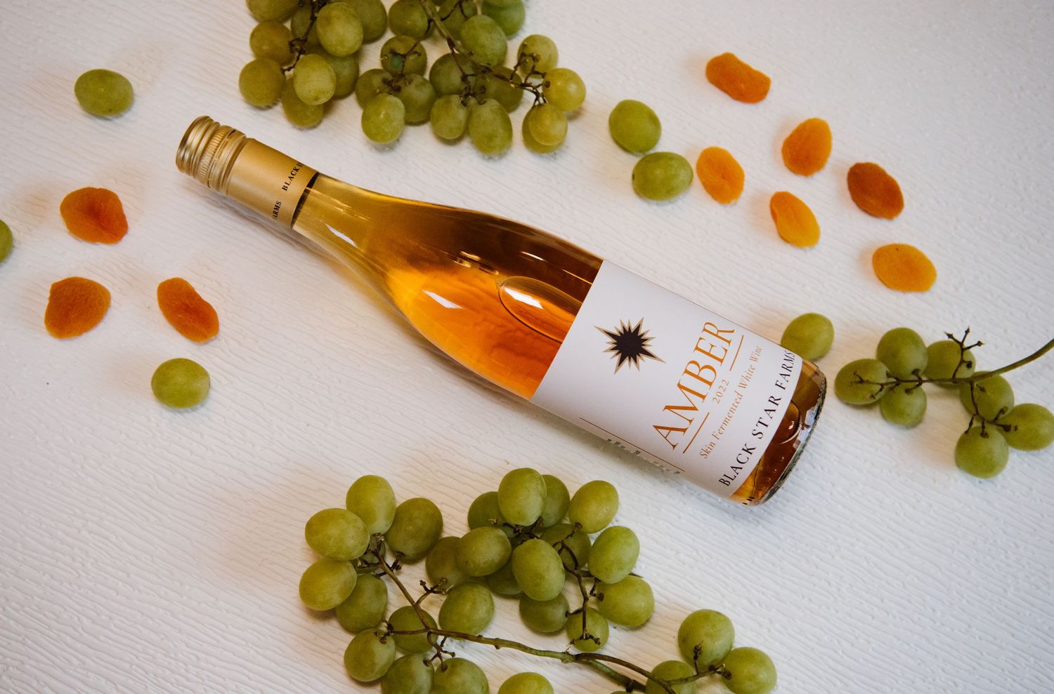 A bottle of golden wine lies on white surface surrounded by fresh green grapes and dried apricots.