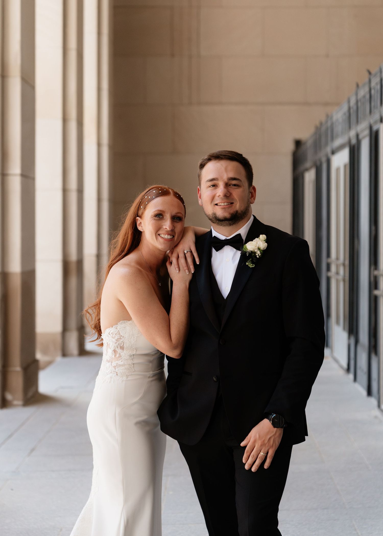 A smiling bride in a white gown and groom in a black tuxedo pose together in an elegant columned hallway.