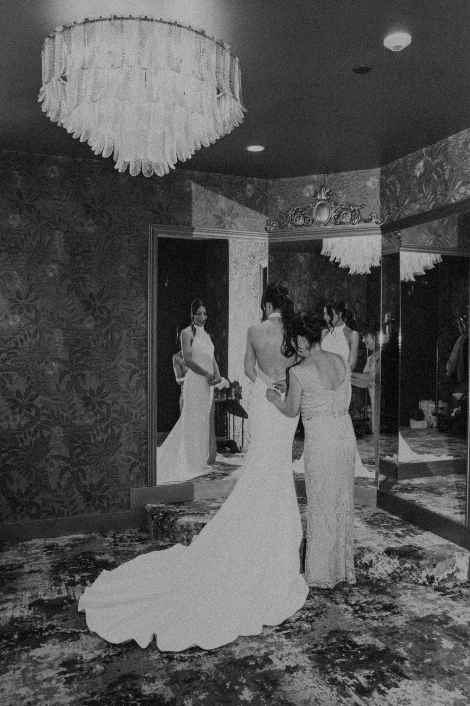 Black and white photo of bride in elegant gown being helped by bridesmaid in ornate mirrored room with chandelier.