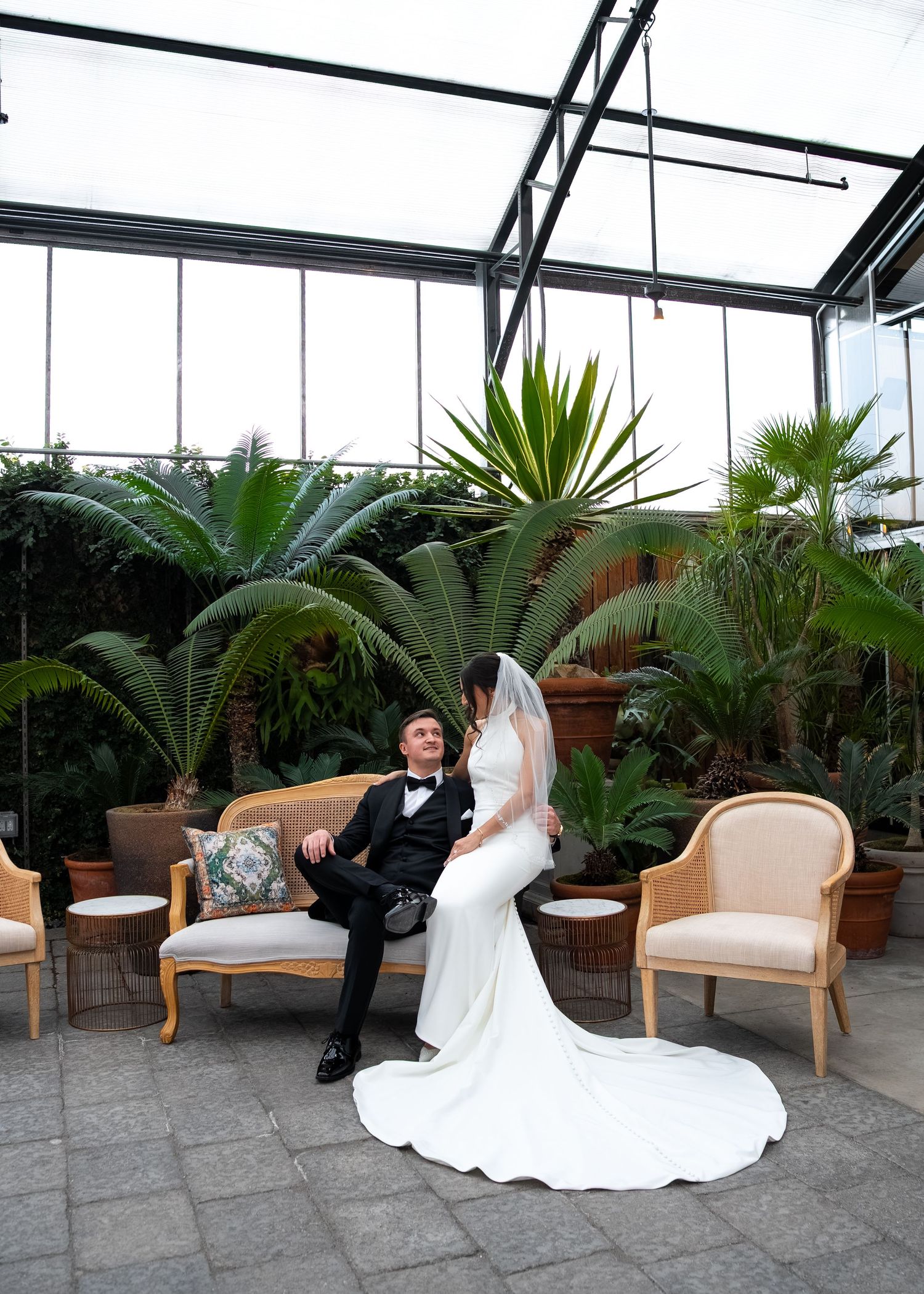 Bride and groom sitting together on an elegant sofa in a glass greenhouse venue surrounded by tropical plants.