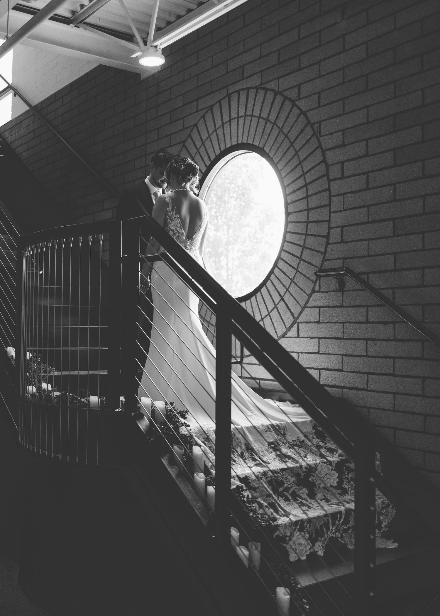 Black and white photo of a dramatic interior staircase with a circular porthole window casting light on brick walls.