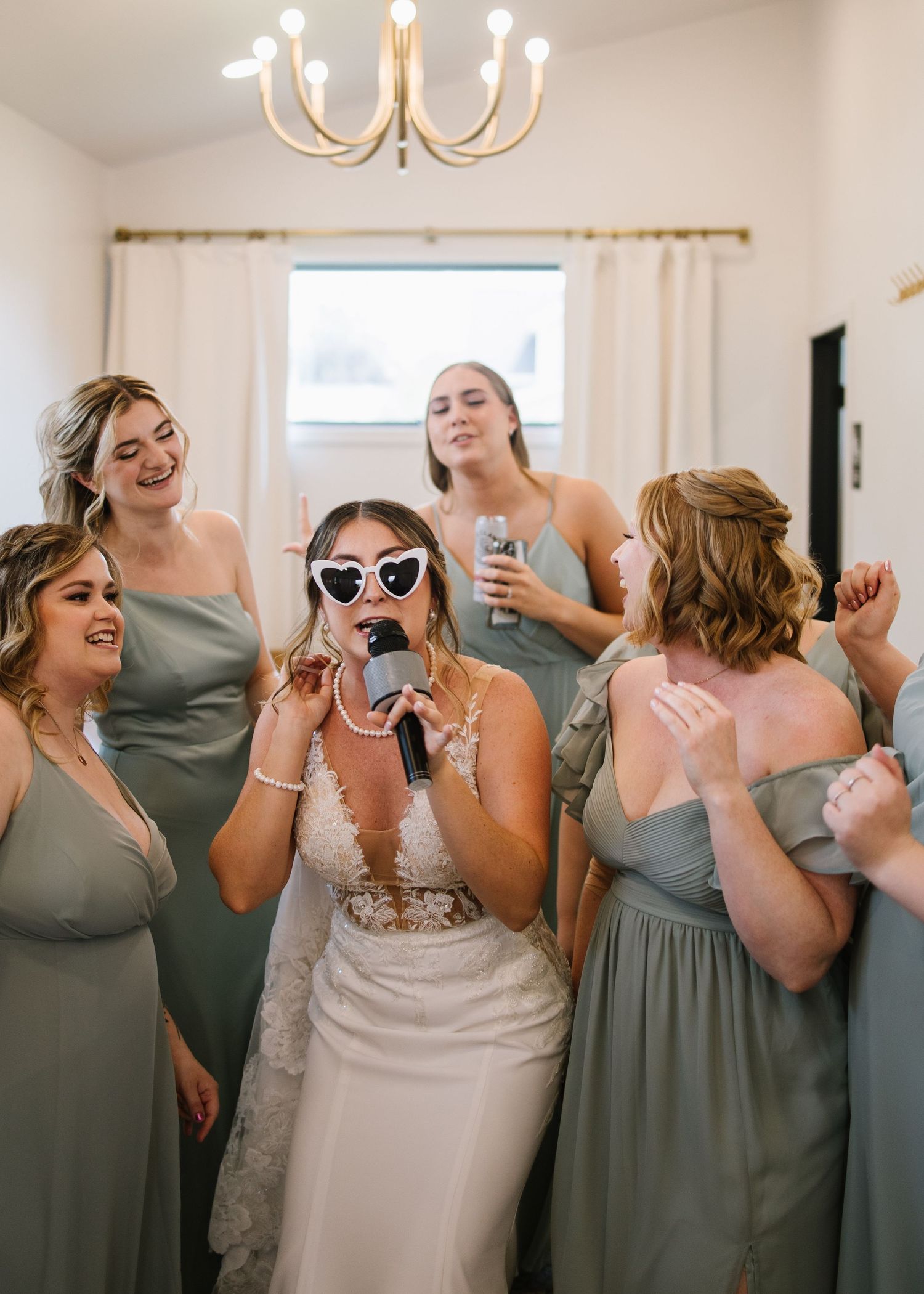 A wedding party in sage green bridesmaid dresses celebrates joyfully together in a light-filled room.