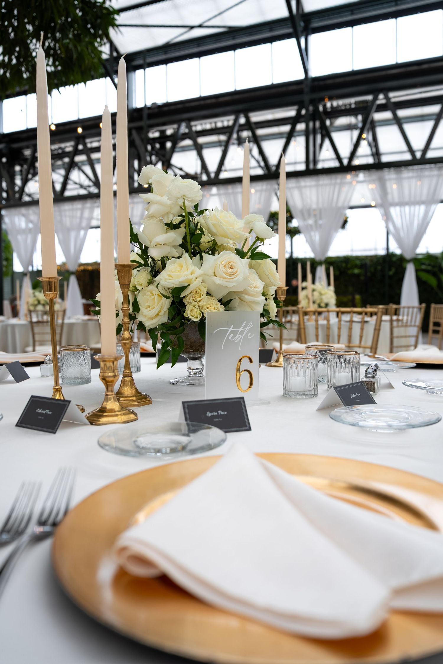Elegant wedding reception table with white roses, gold candlesticks, charger plates, and white napkins in a greenhouse venue.