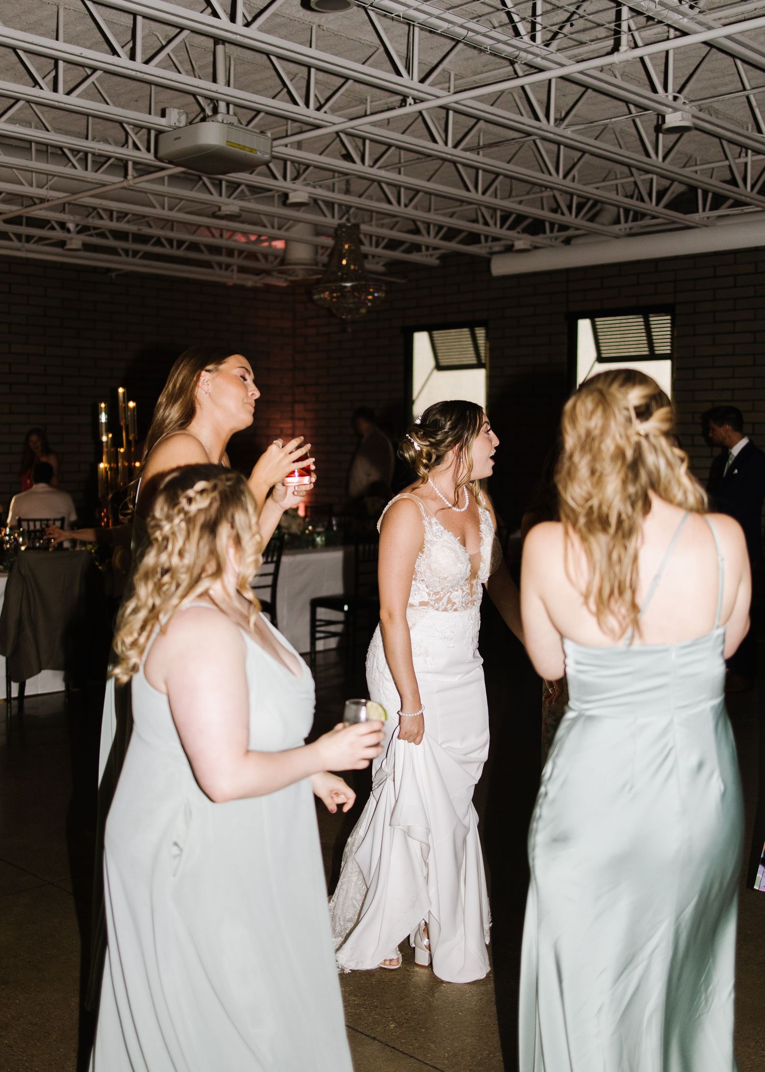 A group dances together at a wedding reception in an industrial-style venue with exposed brick walls.