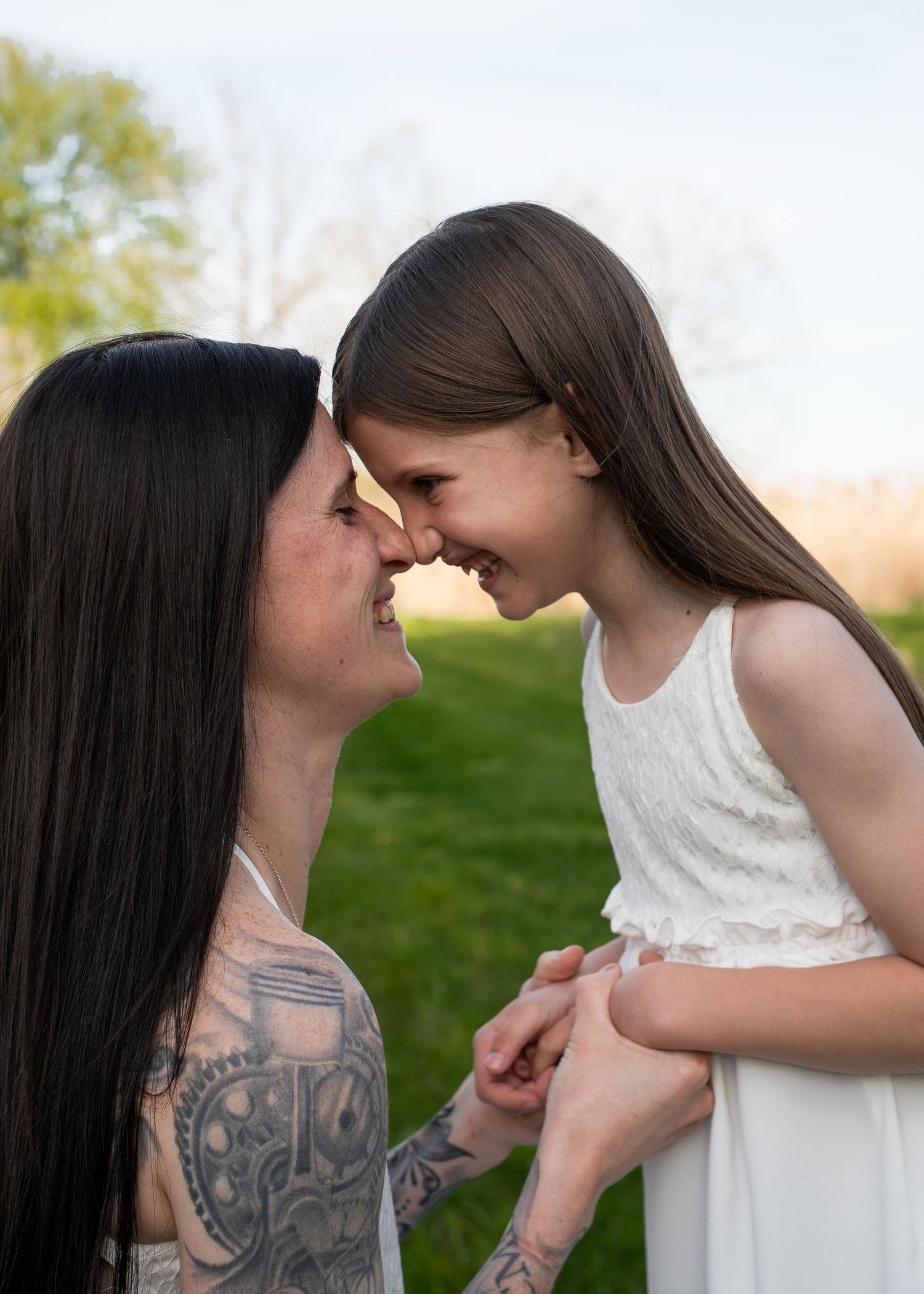 A tattooed mother and young daughter touch foreheads and smile tenderly outdoors in a green park setting.