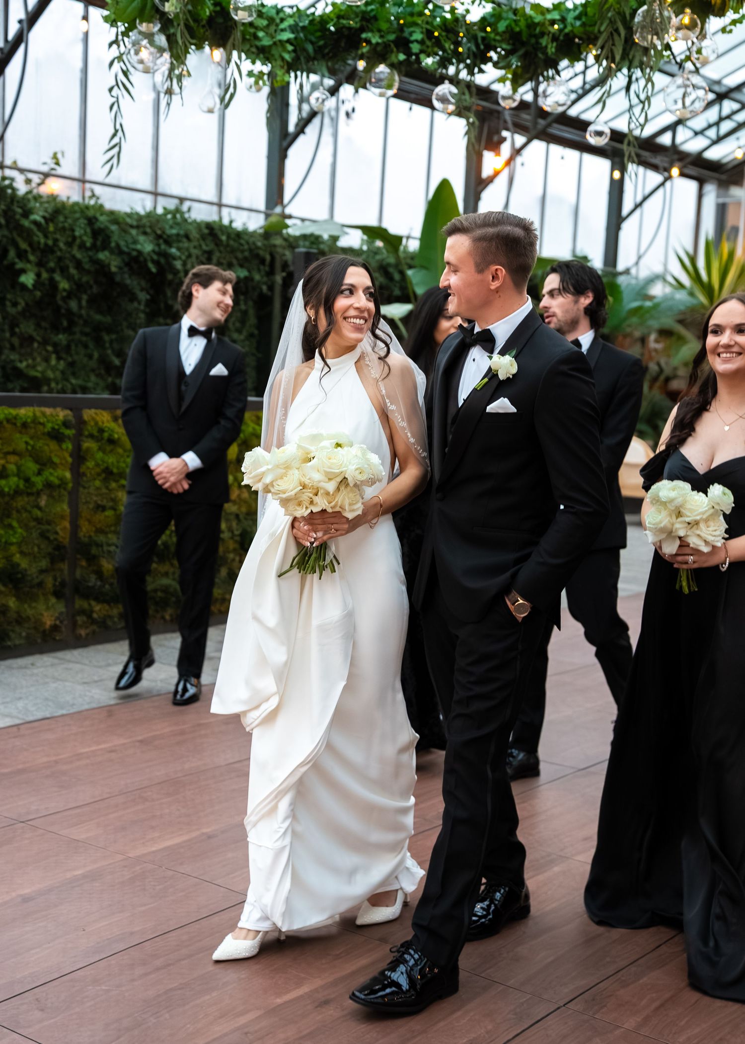A smiling bride in a white gown holds white roses beside her groom in a black tuxedo at an elegant greenhouse wedding.