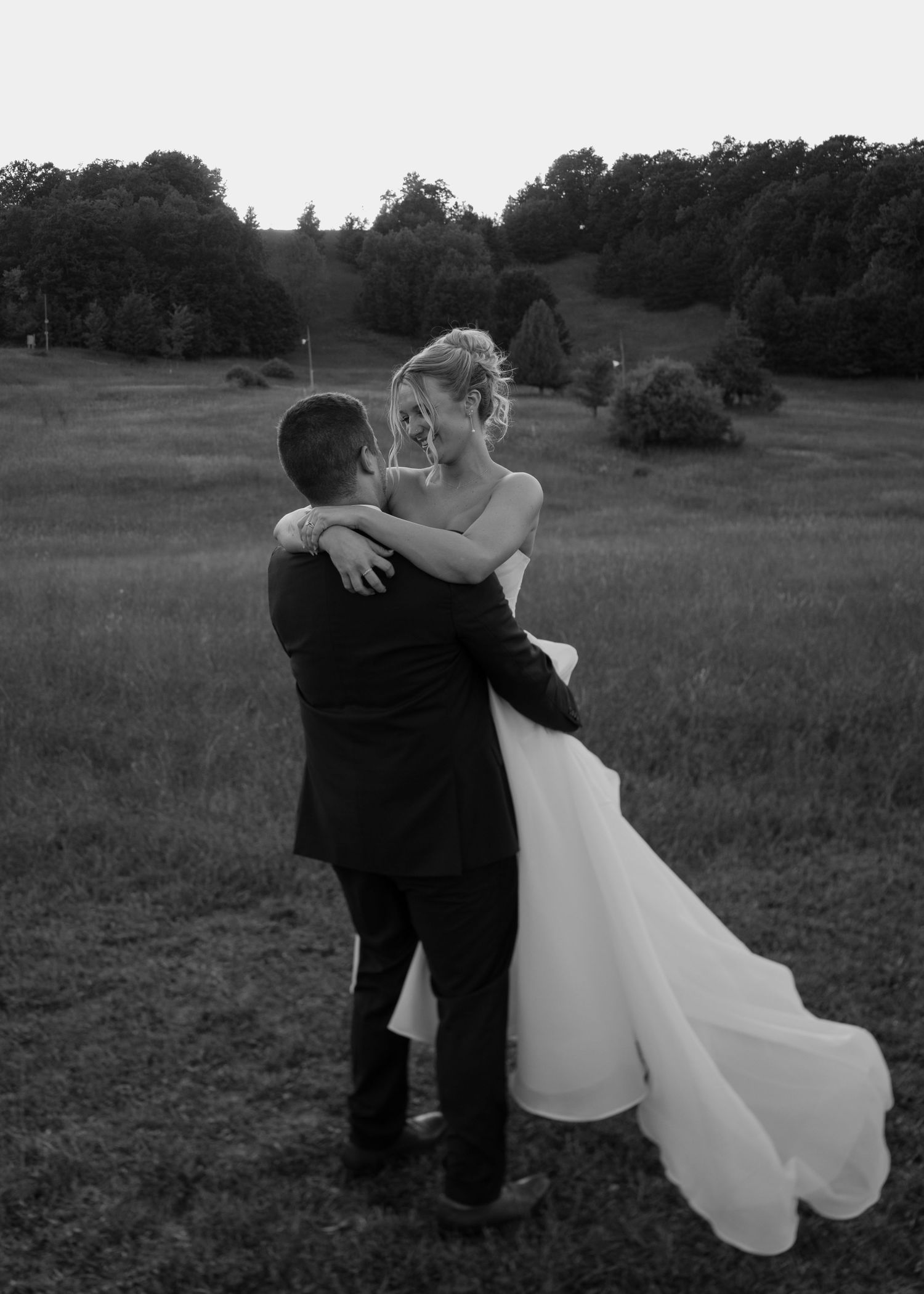 A couple in wedding attire embraces while dancing in a grassy field at sunset.