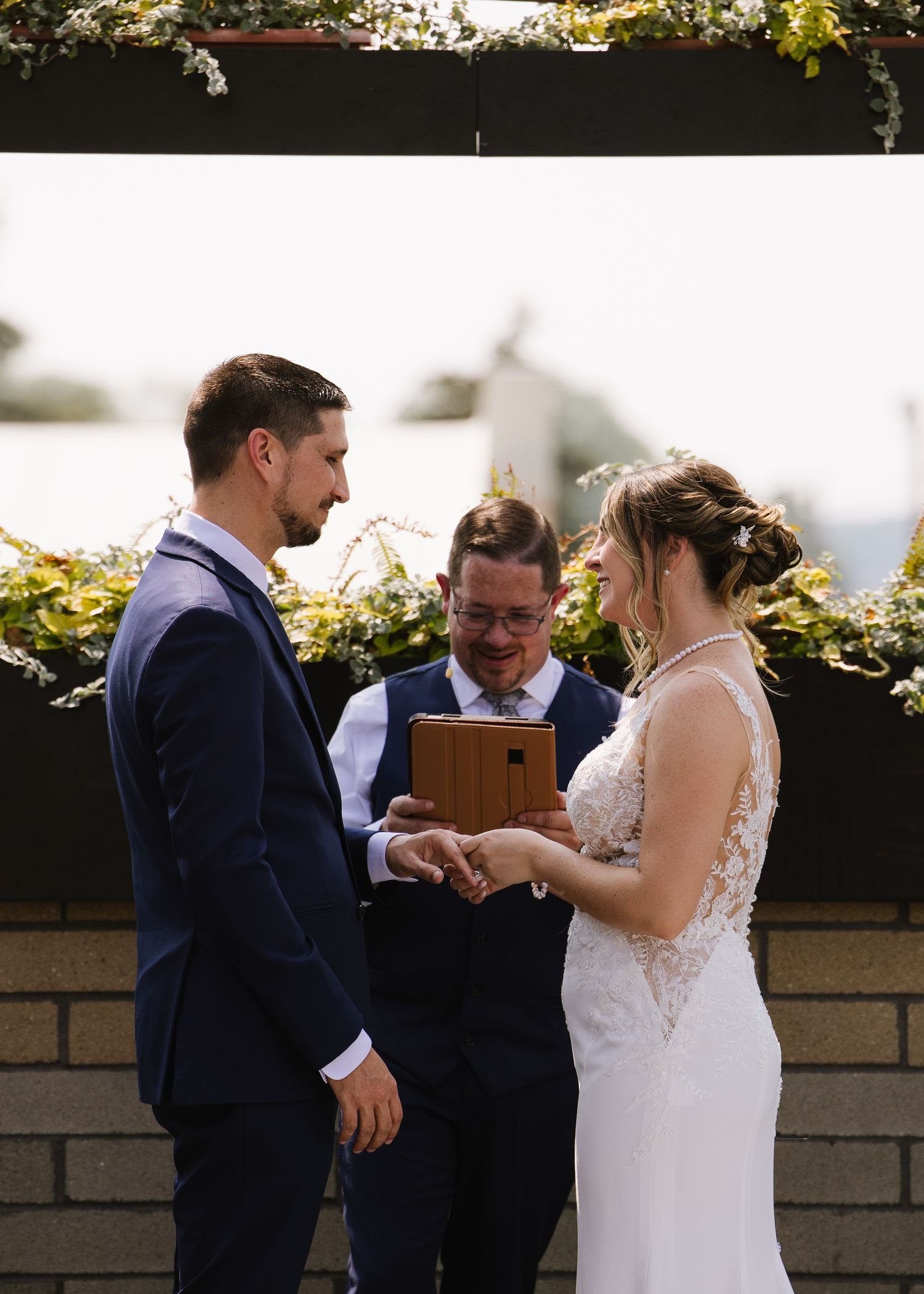 Wedding ceremony takes place outdoors as a couple exchanges rings with officiant on sunny day with floral backdrop.