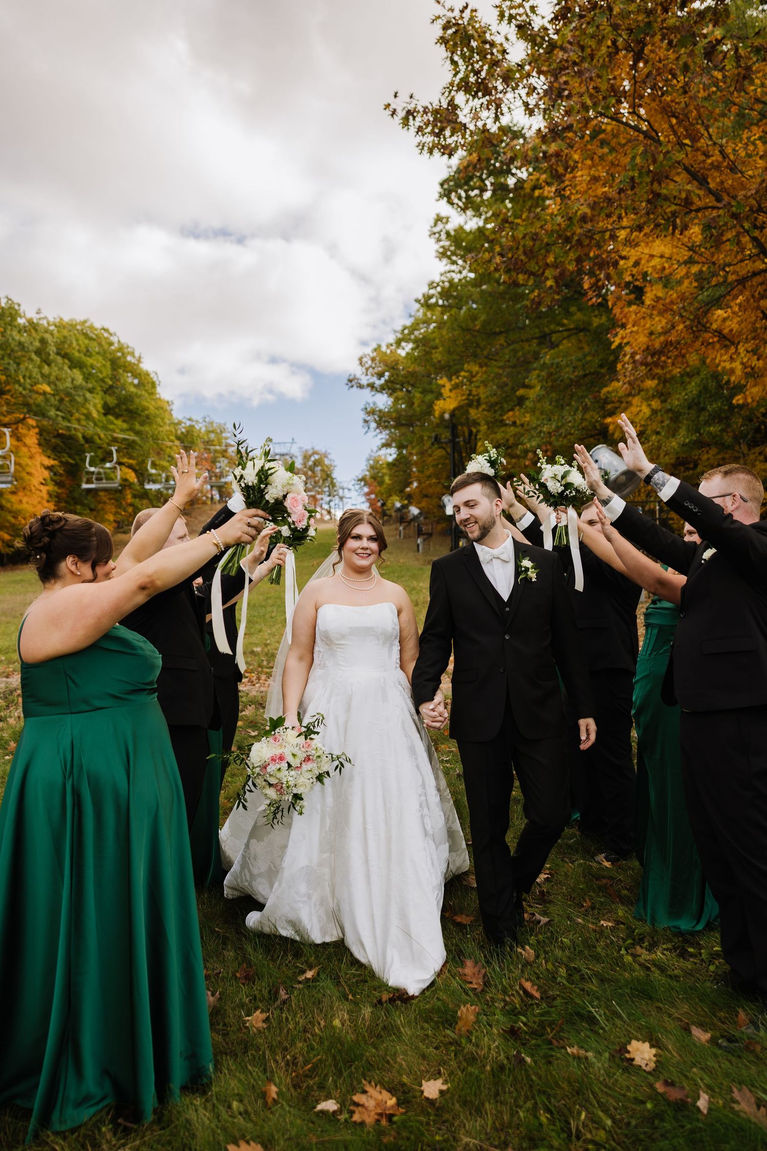 Bride and groom walk through arch of raised bouquets with wedding party in green dresses amid fall foliage.