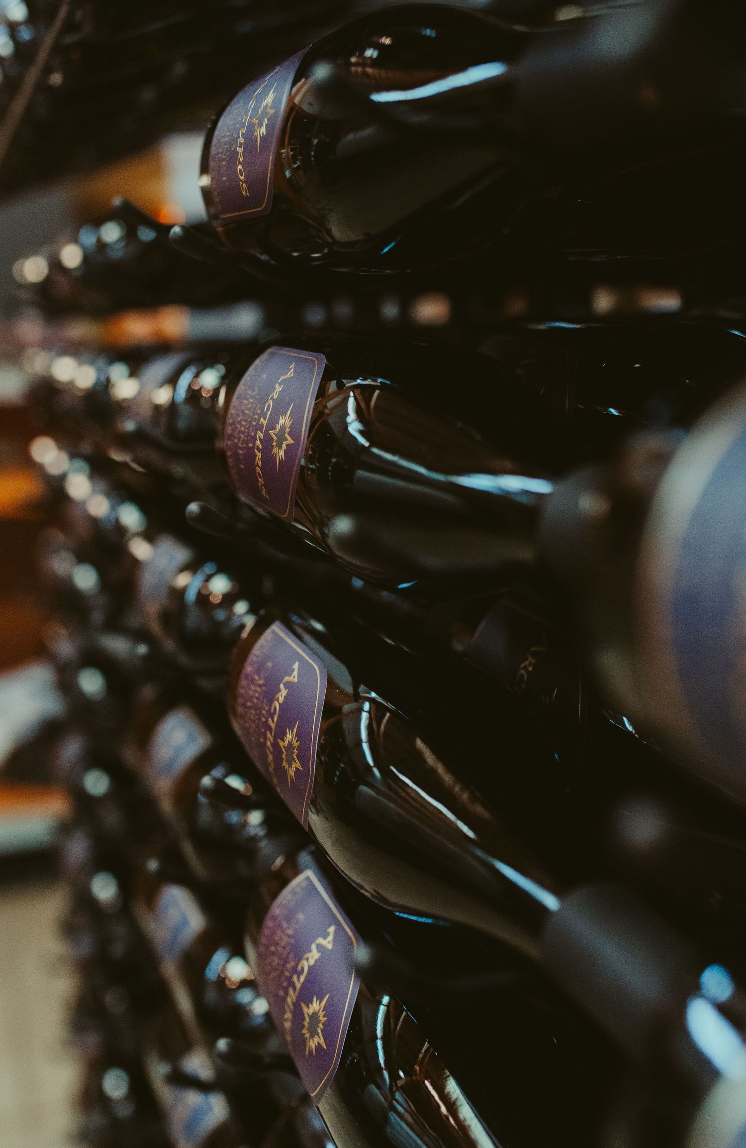 Rows of dark wine bottles with purple labels arranged in a wine cellar rack with moody lighting and shallow depth of field.