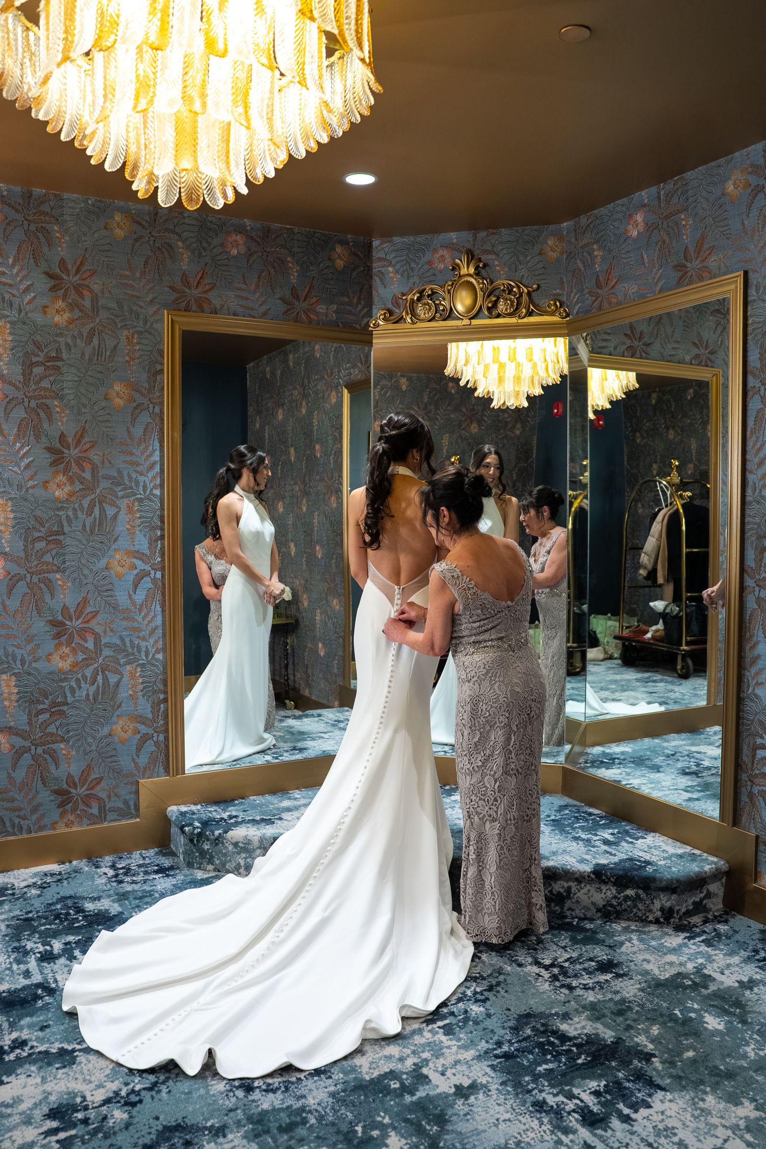 Bride in white gown being helped by bridesmaid in elegant mirrored dressing room with gold chandelier and floral wallpaper.