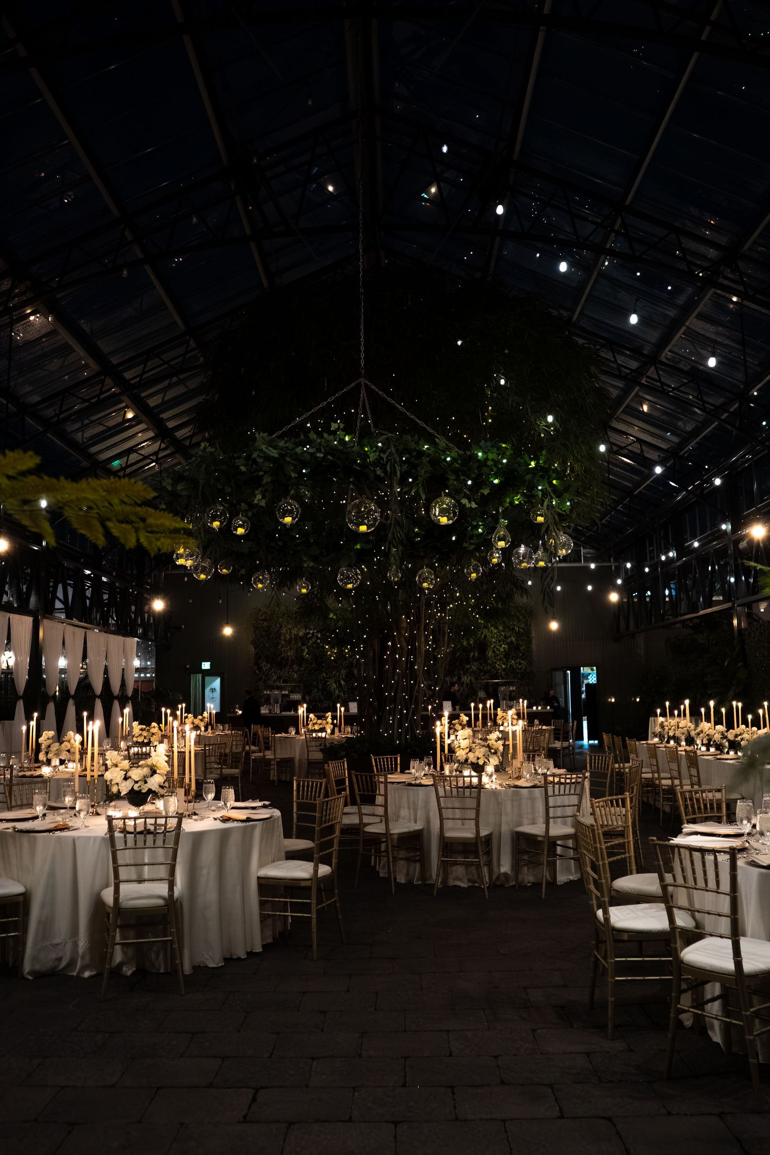 Elegant wedding reception in a greenhouse venue with gold chiavari chairs, candlelit tables, and twinkling lights at night.