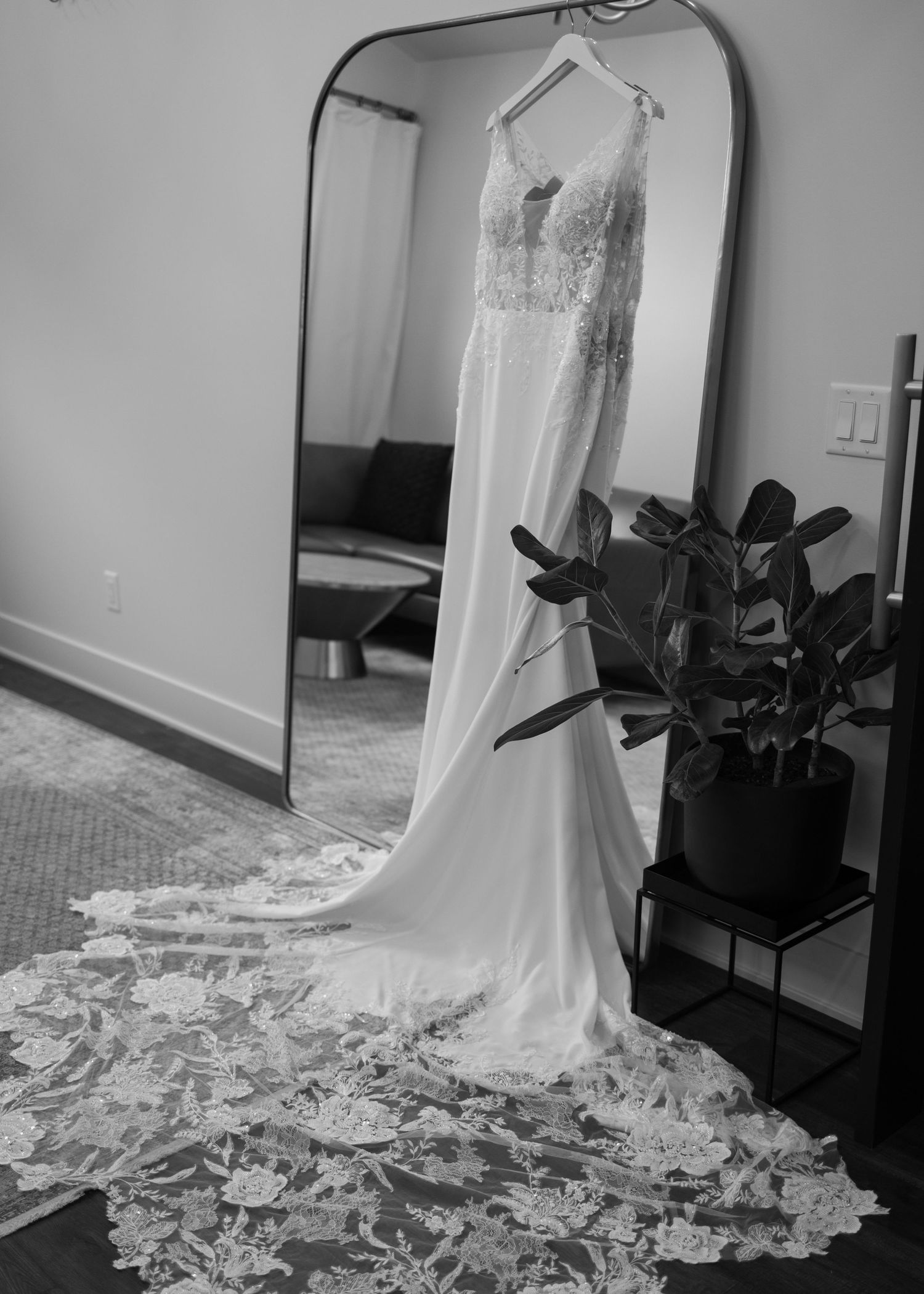 A black and white photo of an elegant wedding dress with lace details and long train hanging in front of a mirror.