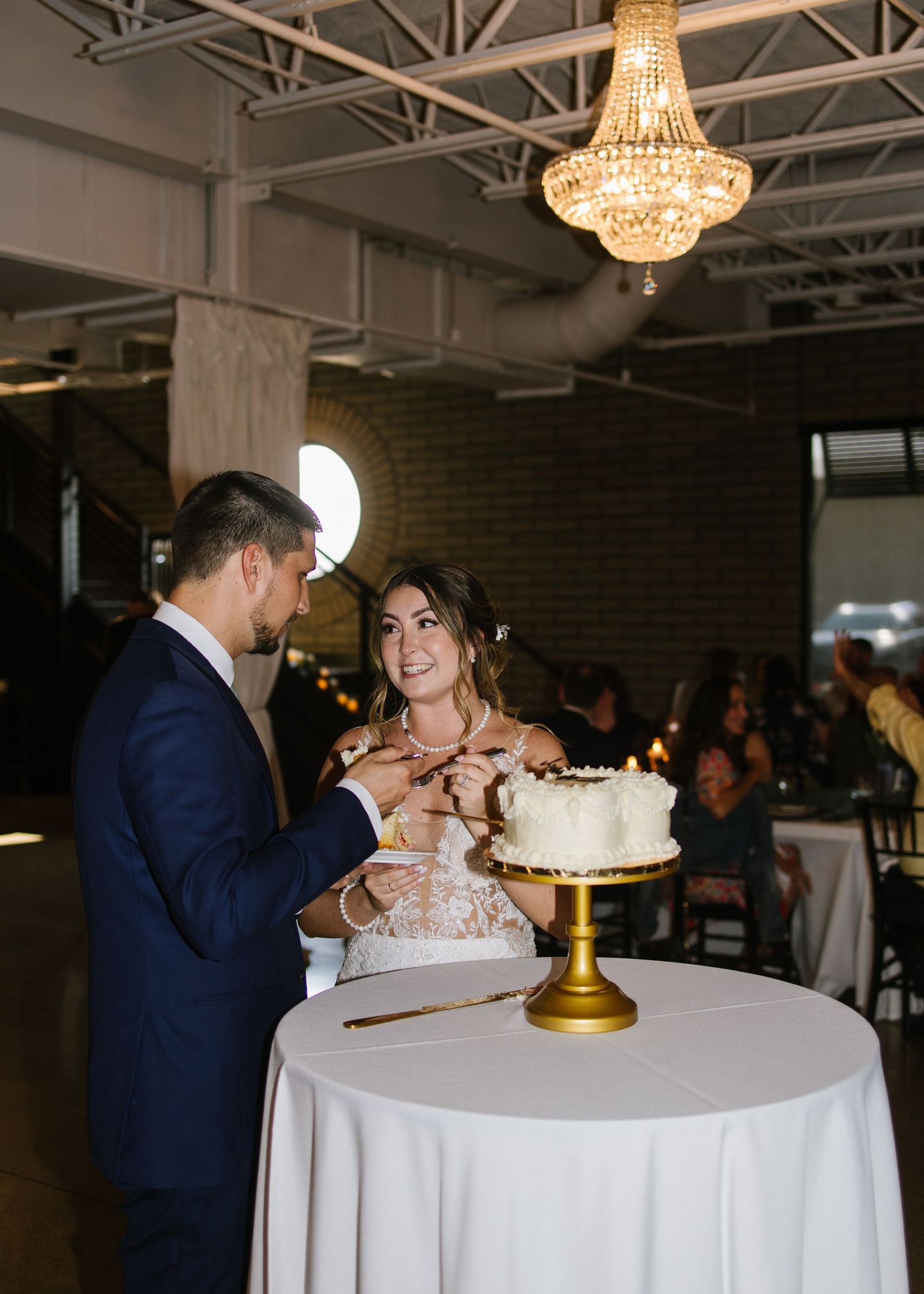 A couple cuts their wedding cake together under a crystal chandelier at an elegant indoor reception venue.