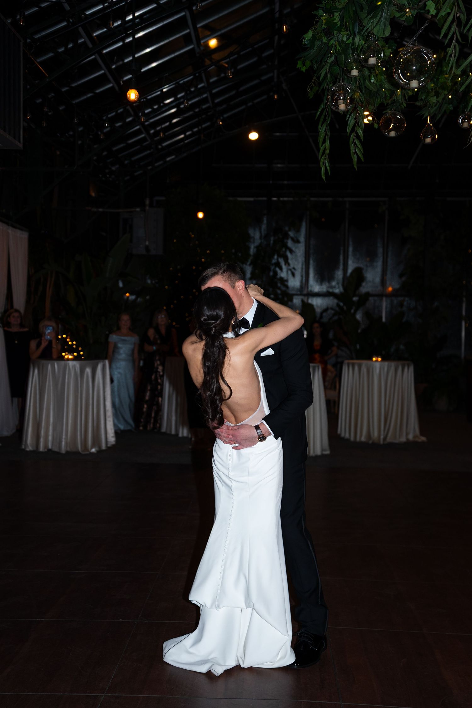 Bride and groom share romantic first dance in dimly lit wedding reception venue with string lights and lush greenery.