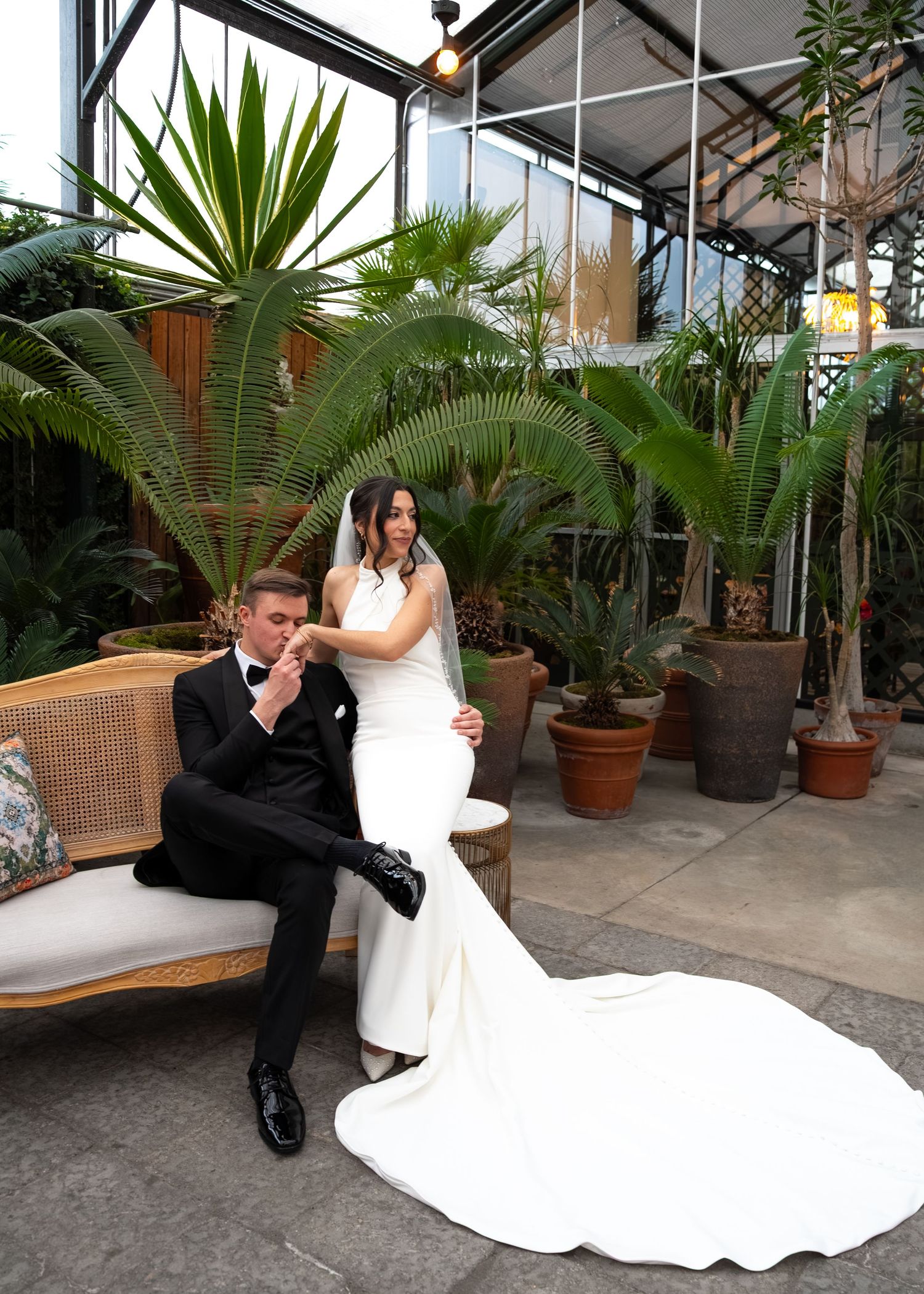 Bride in white mermaid gown and groom in black tuxedo pose in a lush greenhouse venue with tropical plants.