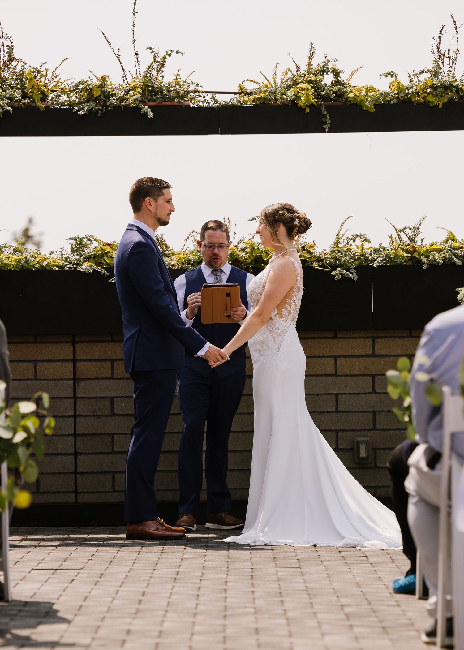 A wedding ceremony takes place on a stone patio with the couple and officiant standing together during the service.