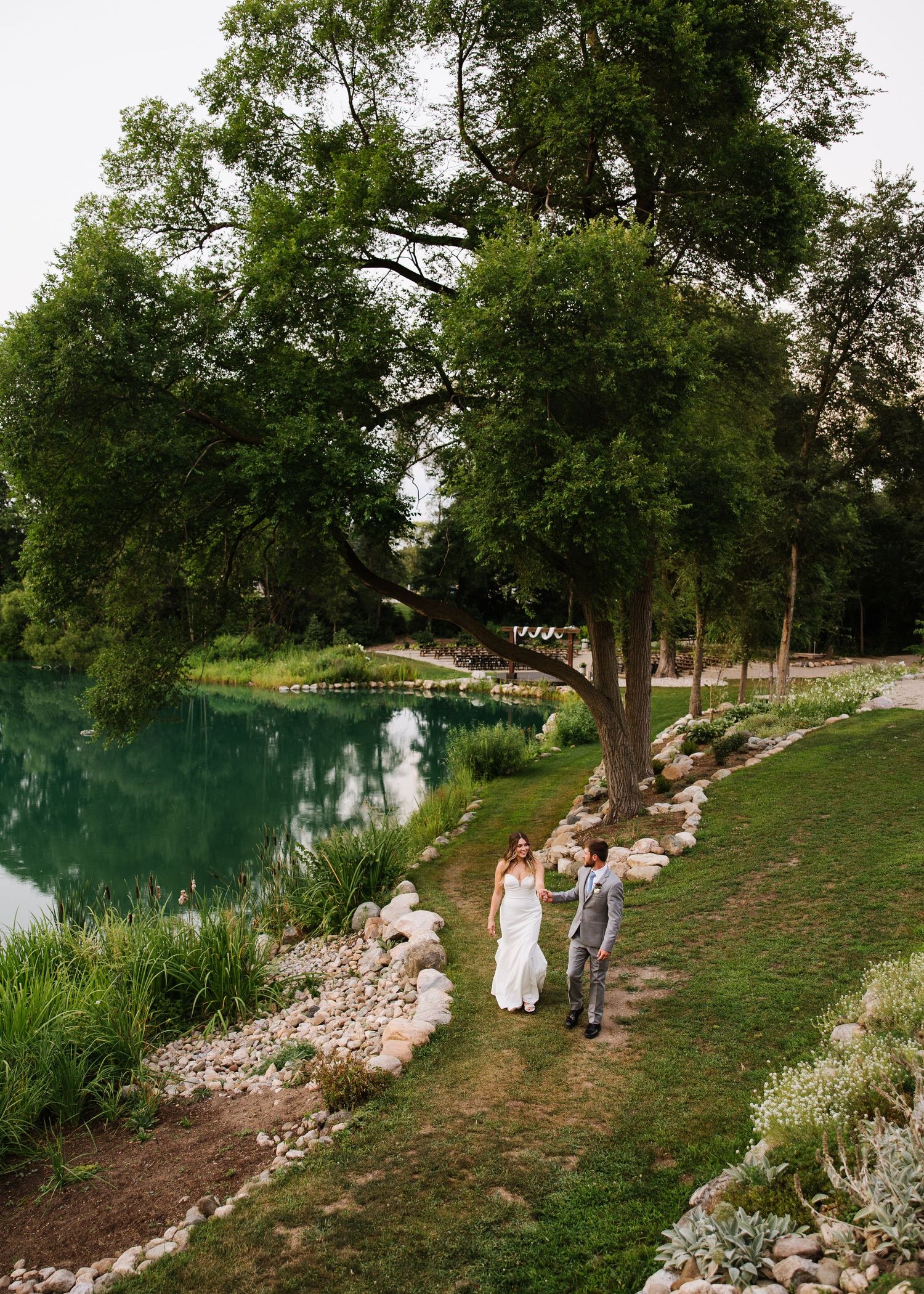 A scenic walking path along a tranquil turquoise pond surrounded by lush trees and rocky landscaping.