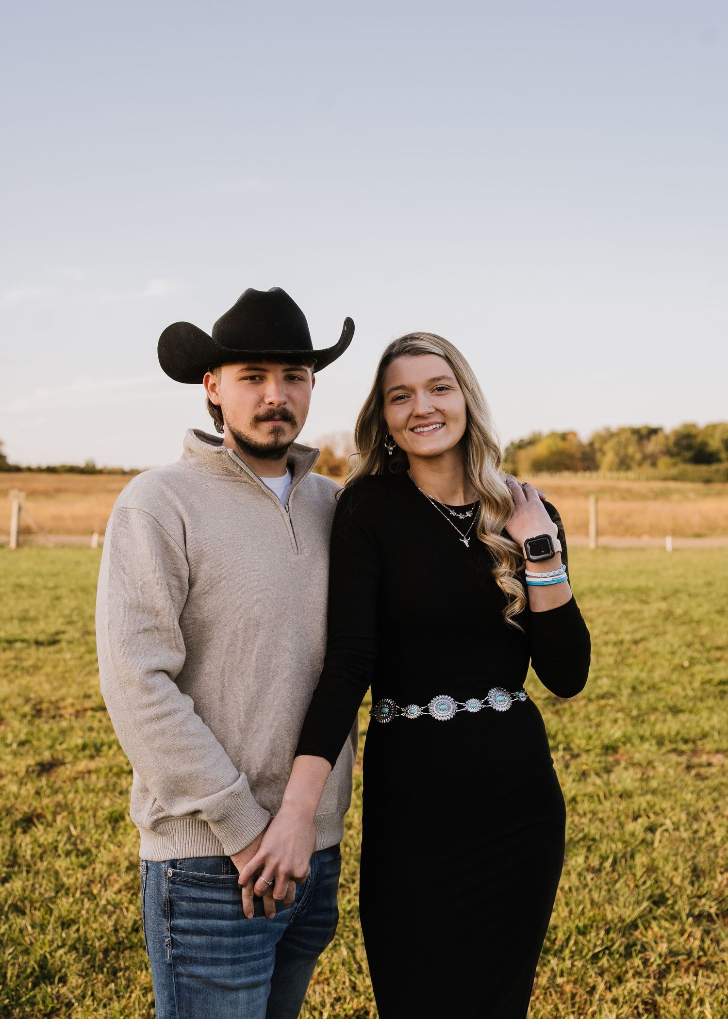 A couple poses together in a grassy field during golden hour wearing western attire and casual clothing.