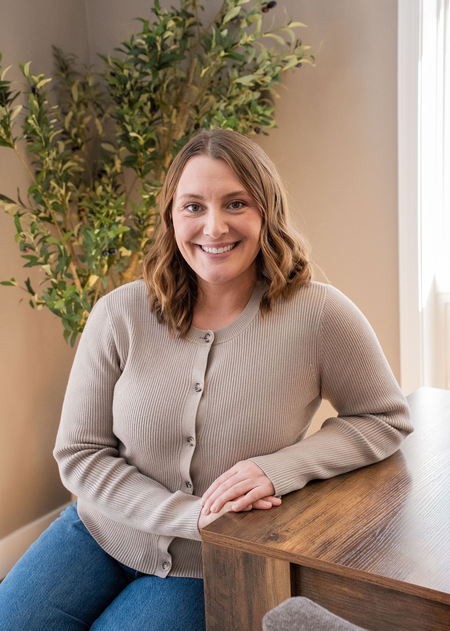 Smiling woman in beige cardigan seated at wooden table with green plant backdrop in bright modern setting.