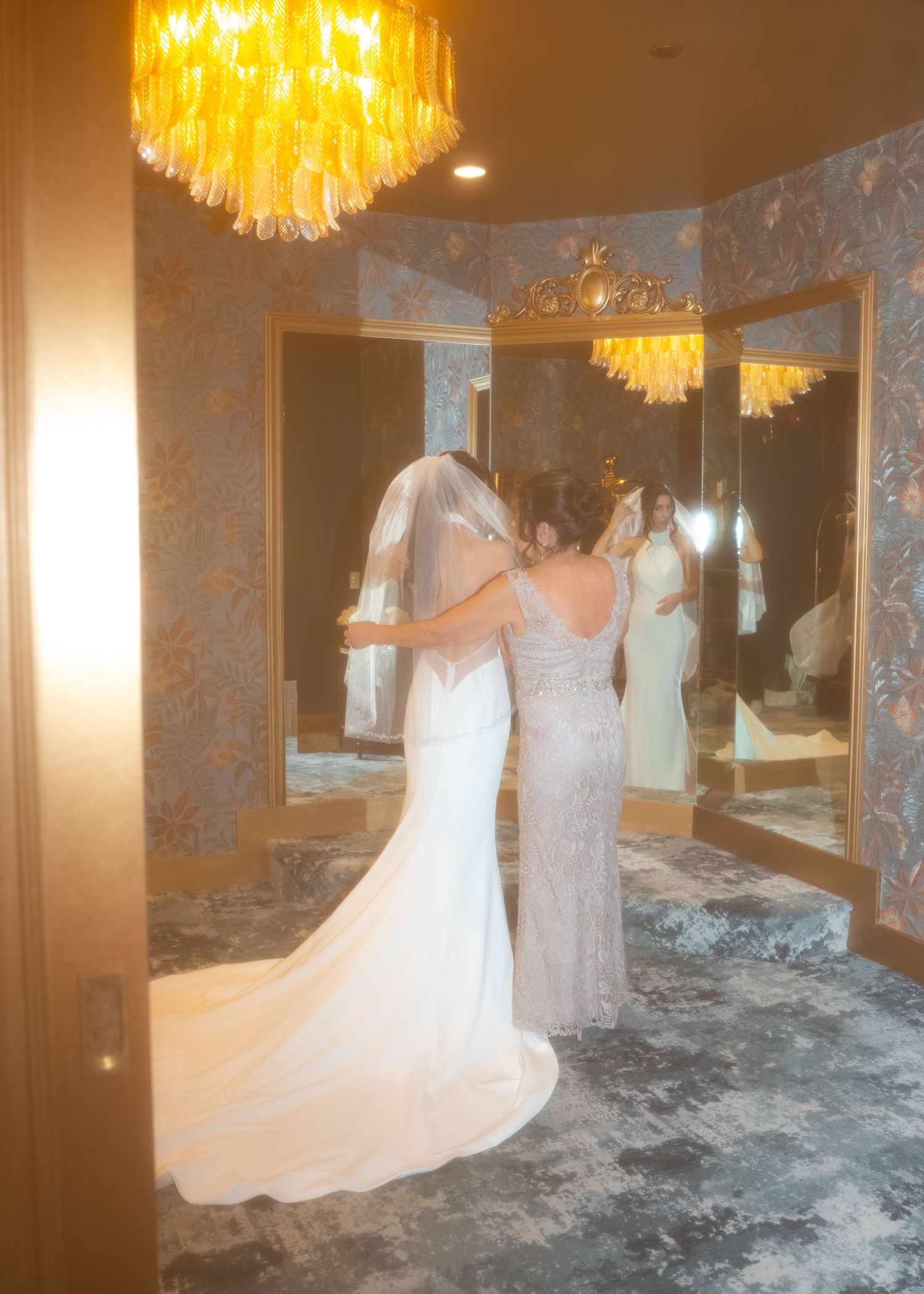 Bride and mother sharing an emotional hug in elegant bridal suite with gold mirrors and chandelier.