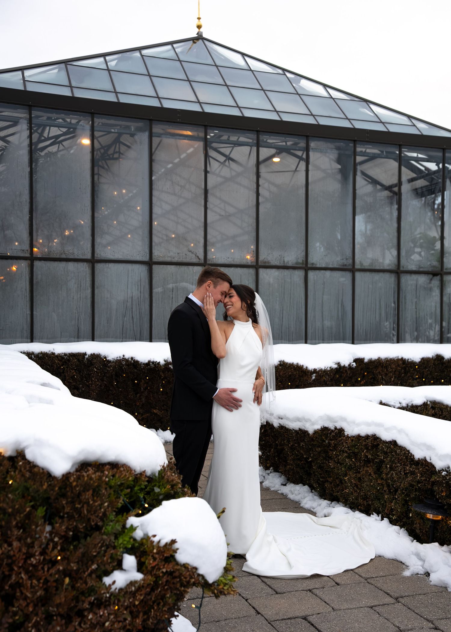 Bride and groom embrace in snow-covered winter garden outside glass greenhouse wedding venue.