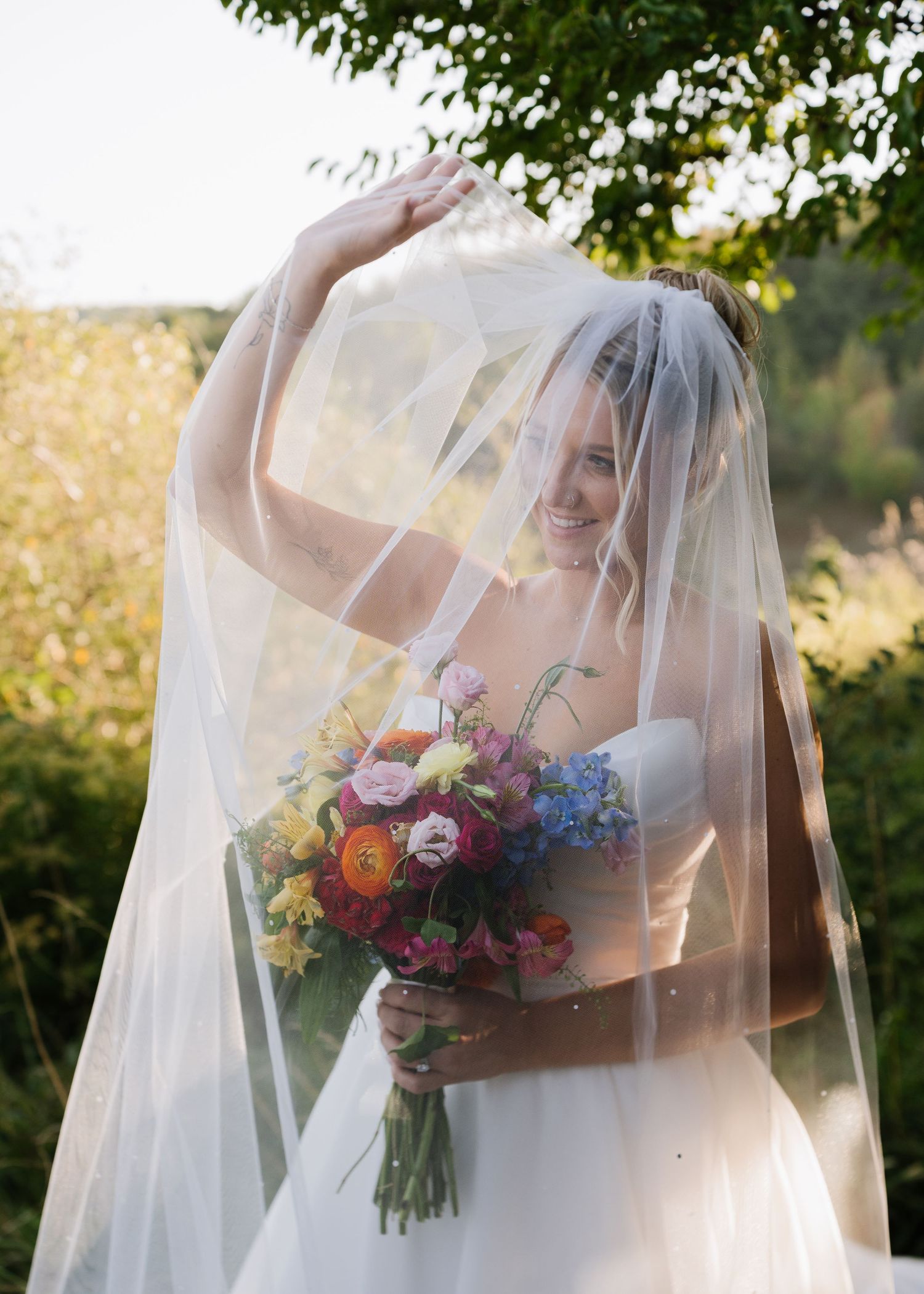 A bride in a floral strapless gown holds a vibrant bouquet while posing under a flowing white veil at sunset.