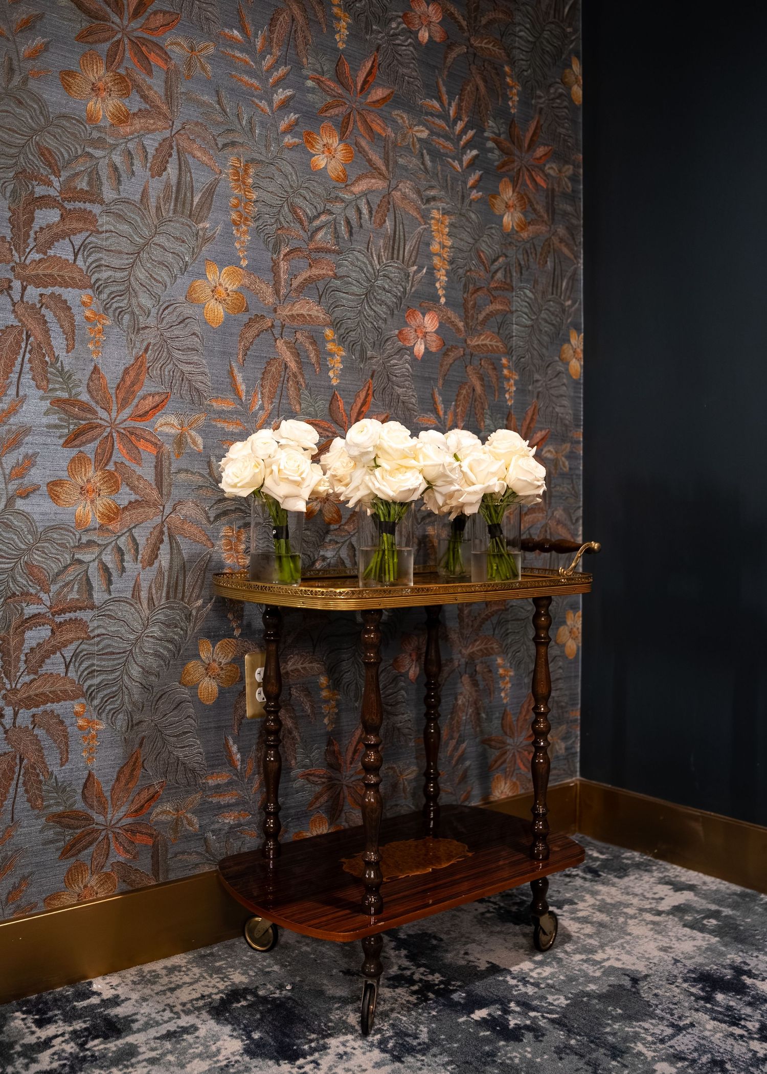 Elegant antique side table with white roses in glass vases against ornate blue and copper tropical floral wallpaper.