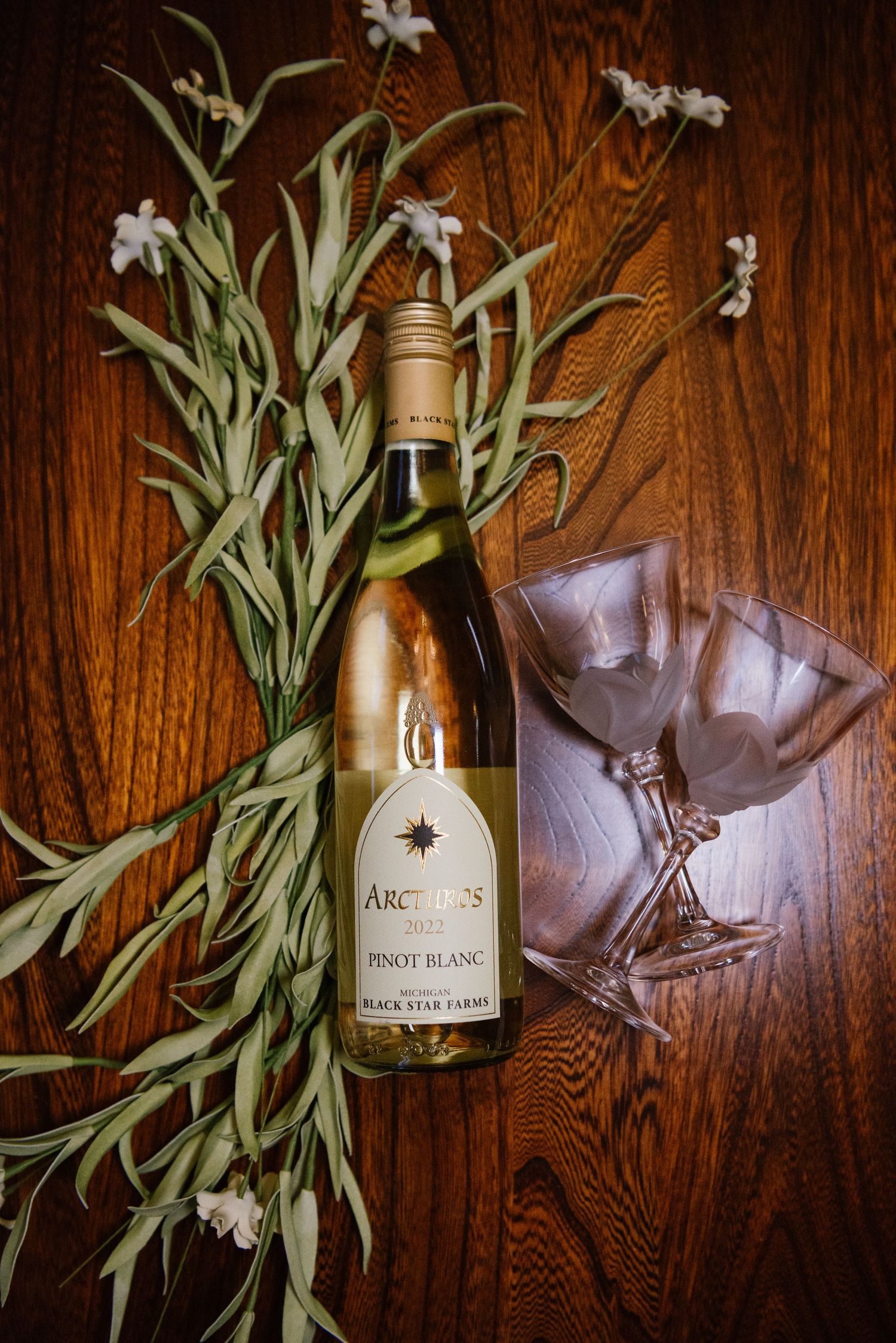 Bottle of Pinot Blanc wine with wine glass on wooden surface decorated with delicate white flowers and green foliage.