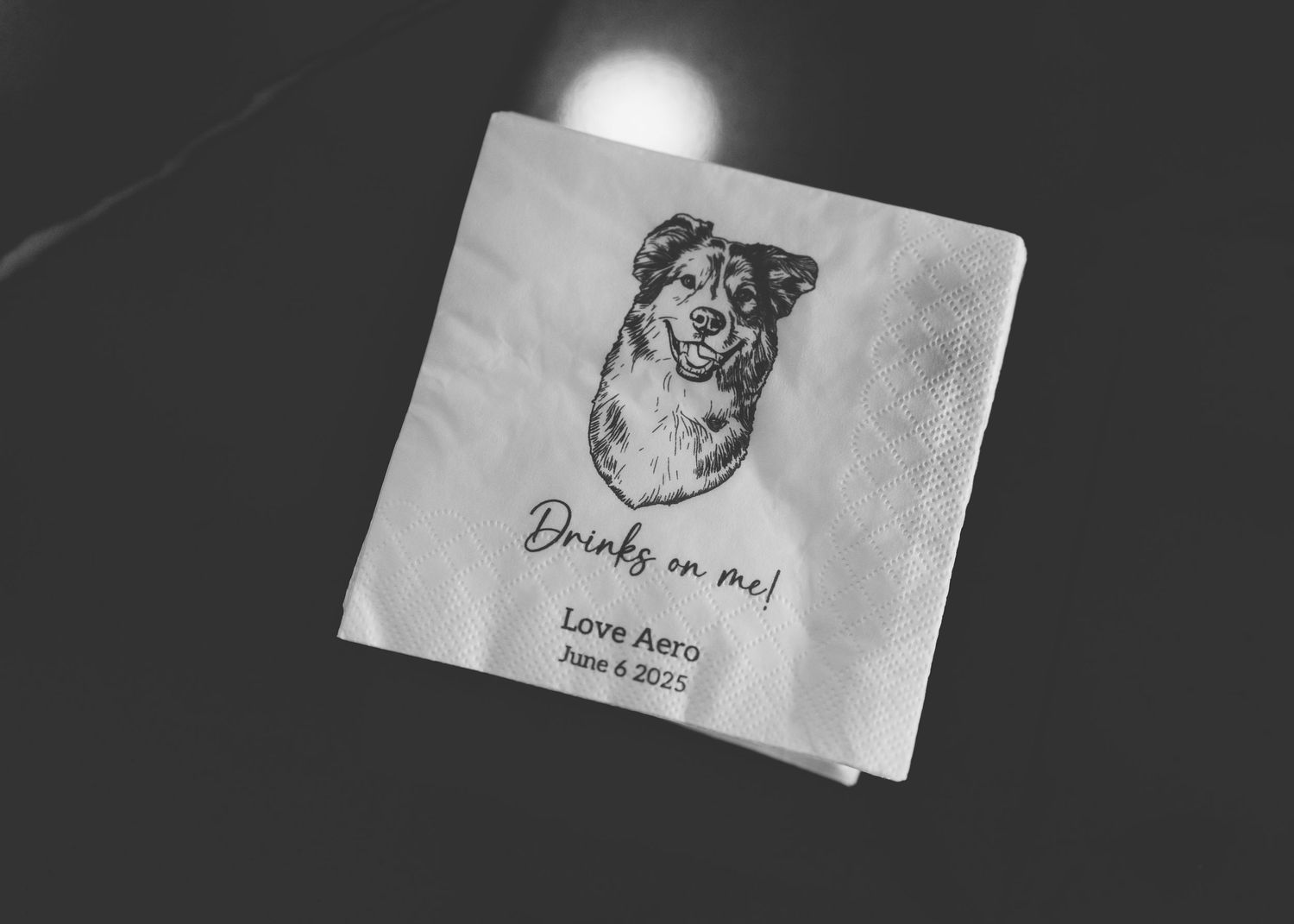 A black and white napkin with a hand-drawn dog illustration and text that says 'Drinks on me!' on a dark surface.