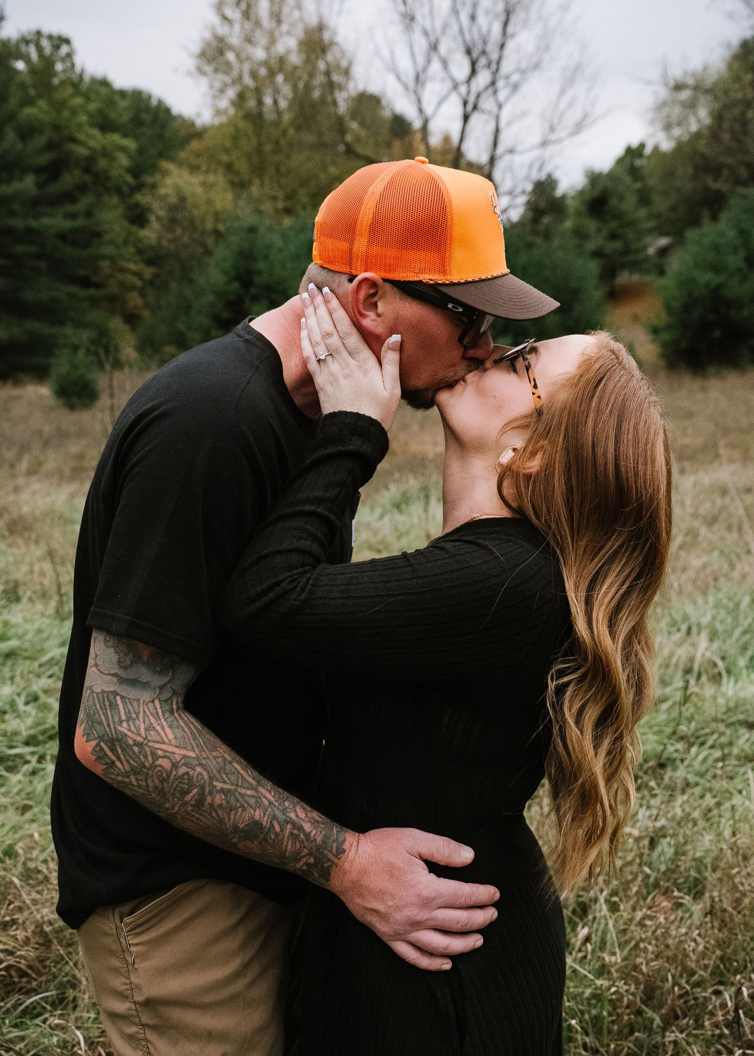 A couple shares a romantic embrace in a field during autumn while one person wears an orange hunting cap.