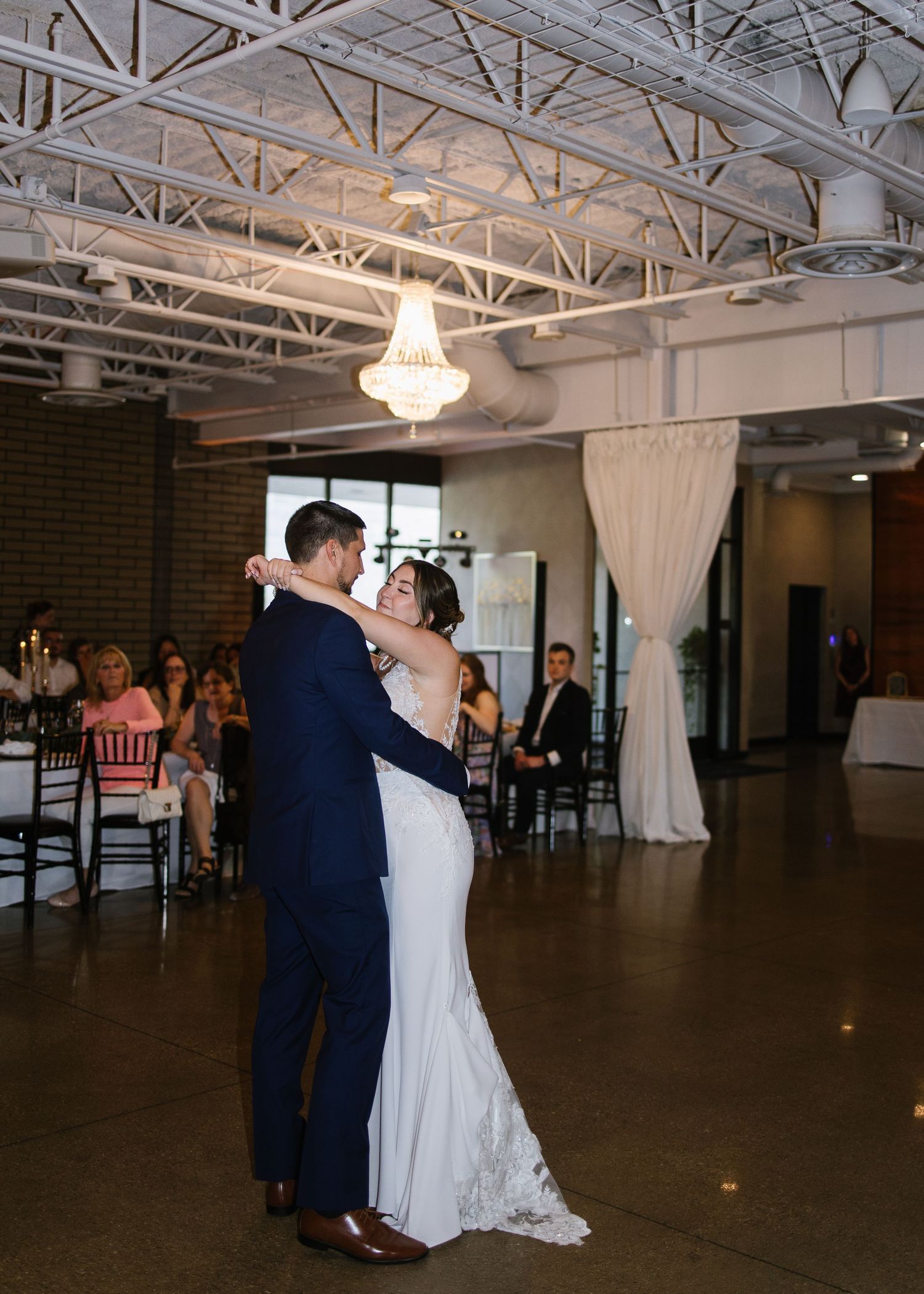 A couple dances together in an industrial-style venue with exposed ceilings and elegant chandeliers.