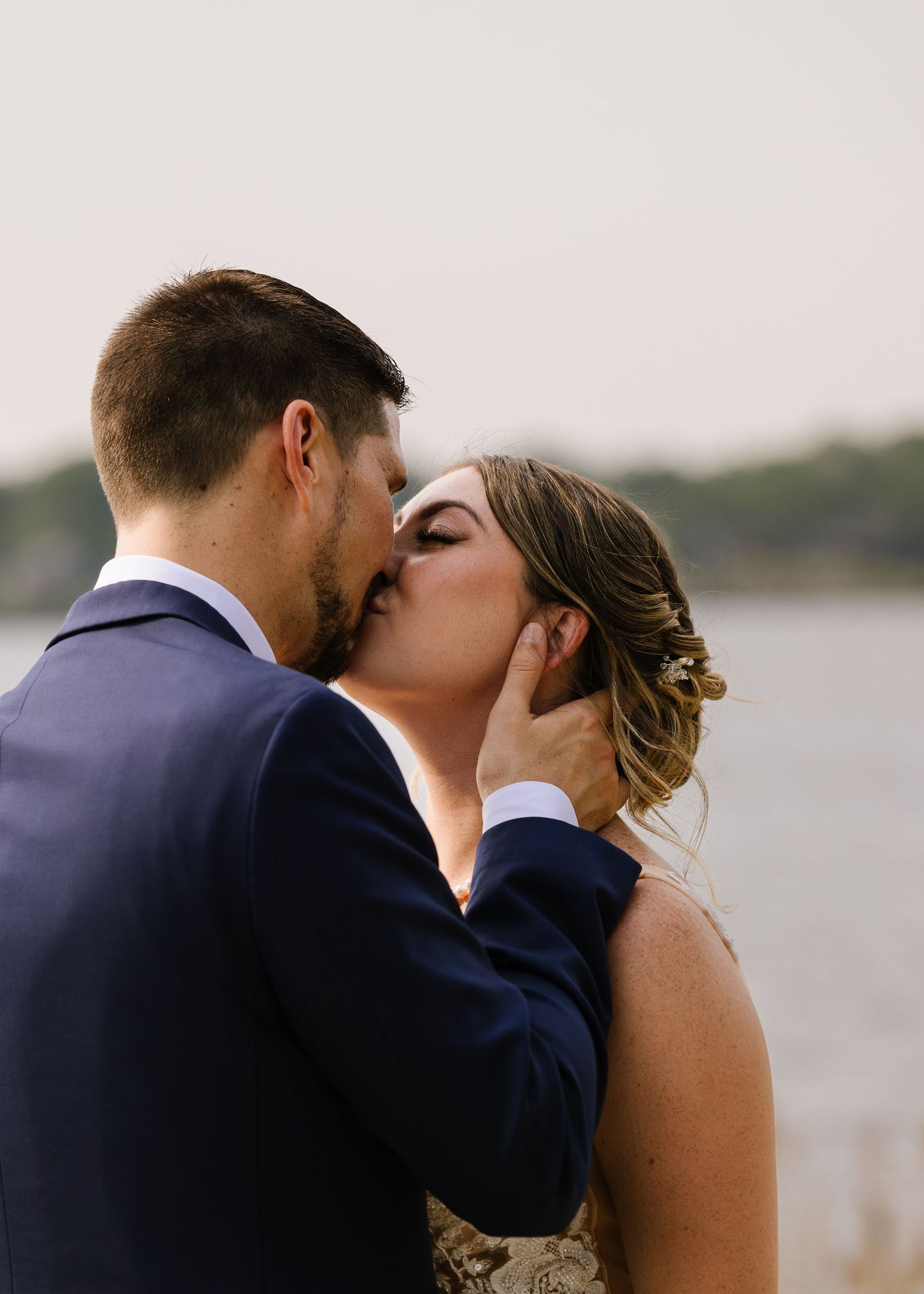 Romantic couple in navy suit and sleeveless dress share multiple tender kisses by a waterfront at sunset.
