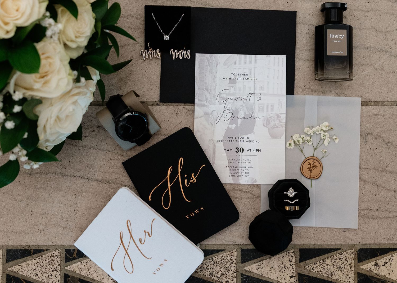 Elegant wedding flat lay with black and white invitations, His and Hers vow books, ring box, watch, perfume, and white roses.