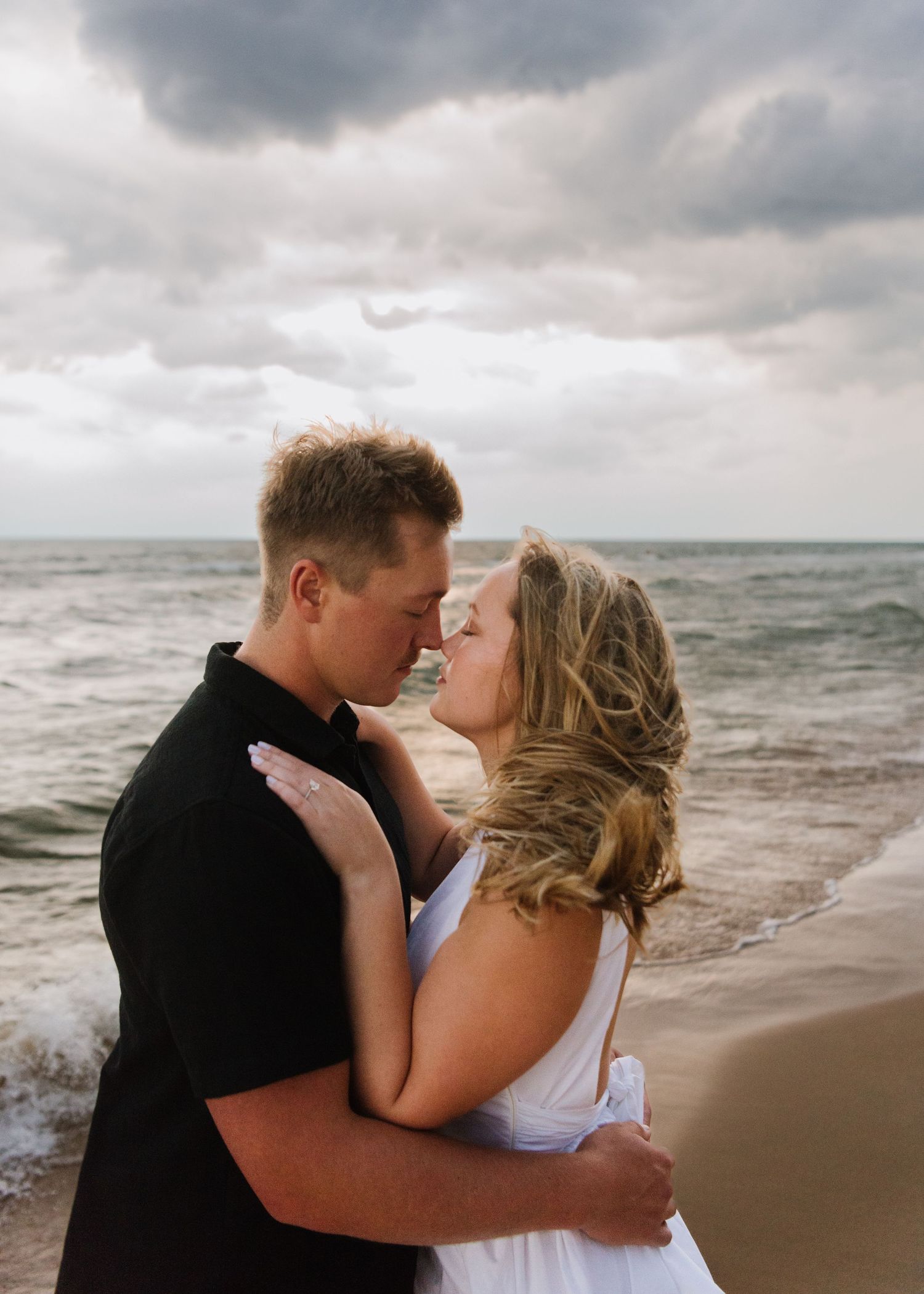 A romantic couple embraces on a moody beach with stormy clouds and crashing waves in the background.