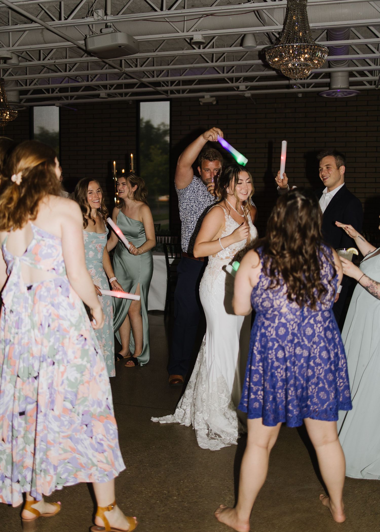 Wedding guests dance and celebrate on the dance floor at an indoor reception venue with elegant chandeliers.