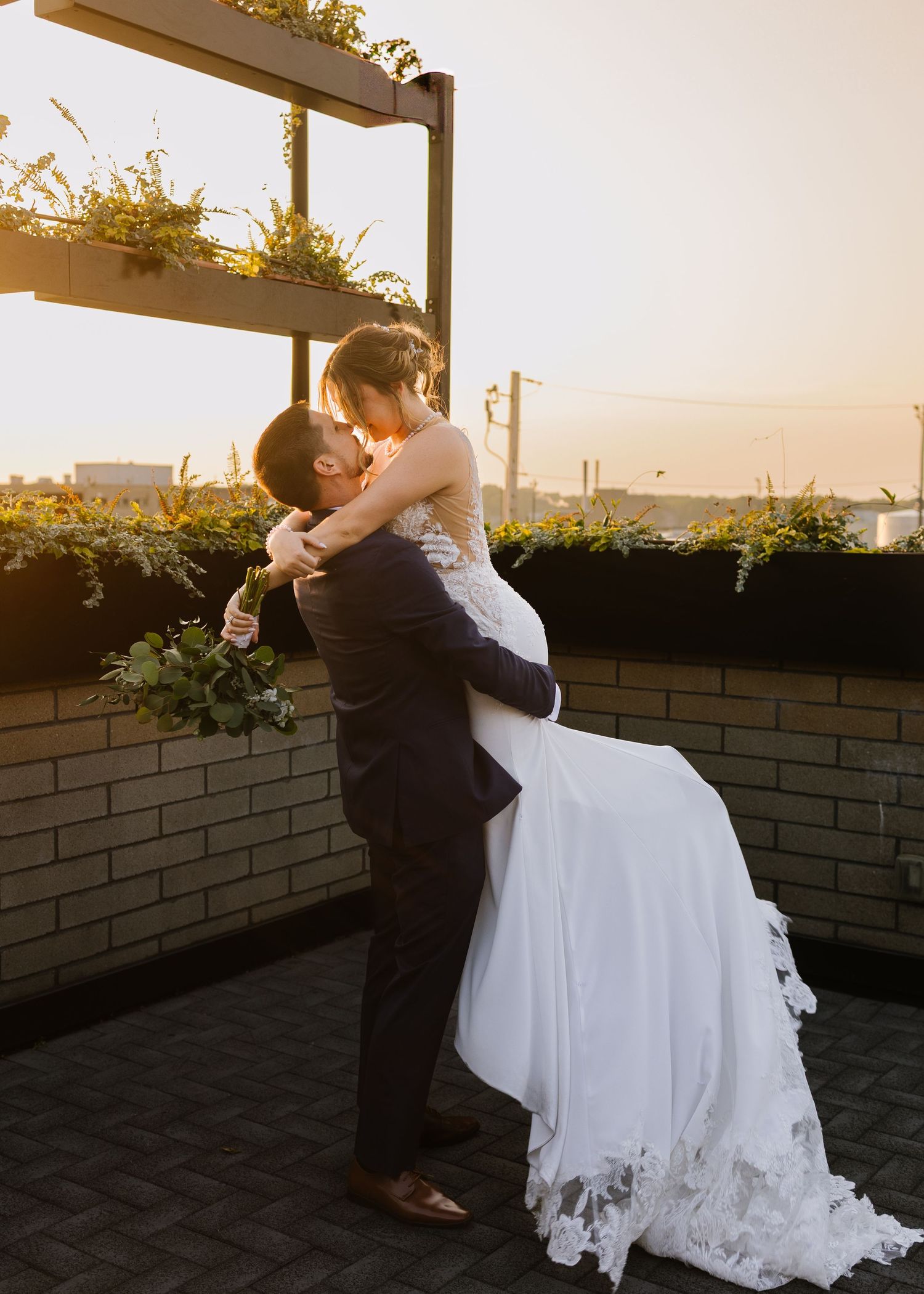 A couple in formal wedding attire share an intimate embrace at sunset on a rooftop terrace with climbing vines and brick walls.
