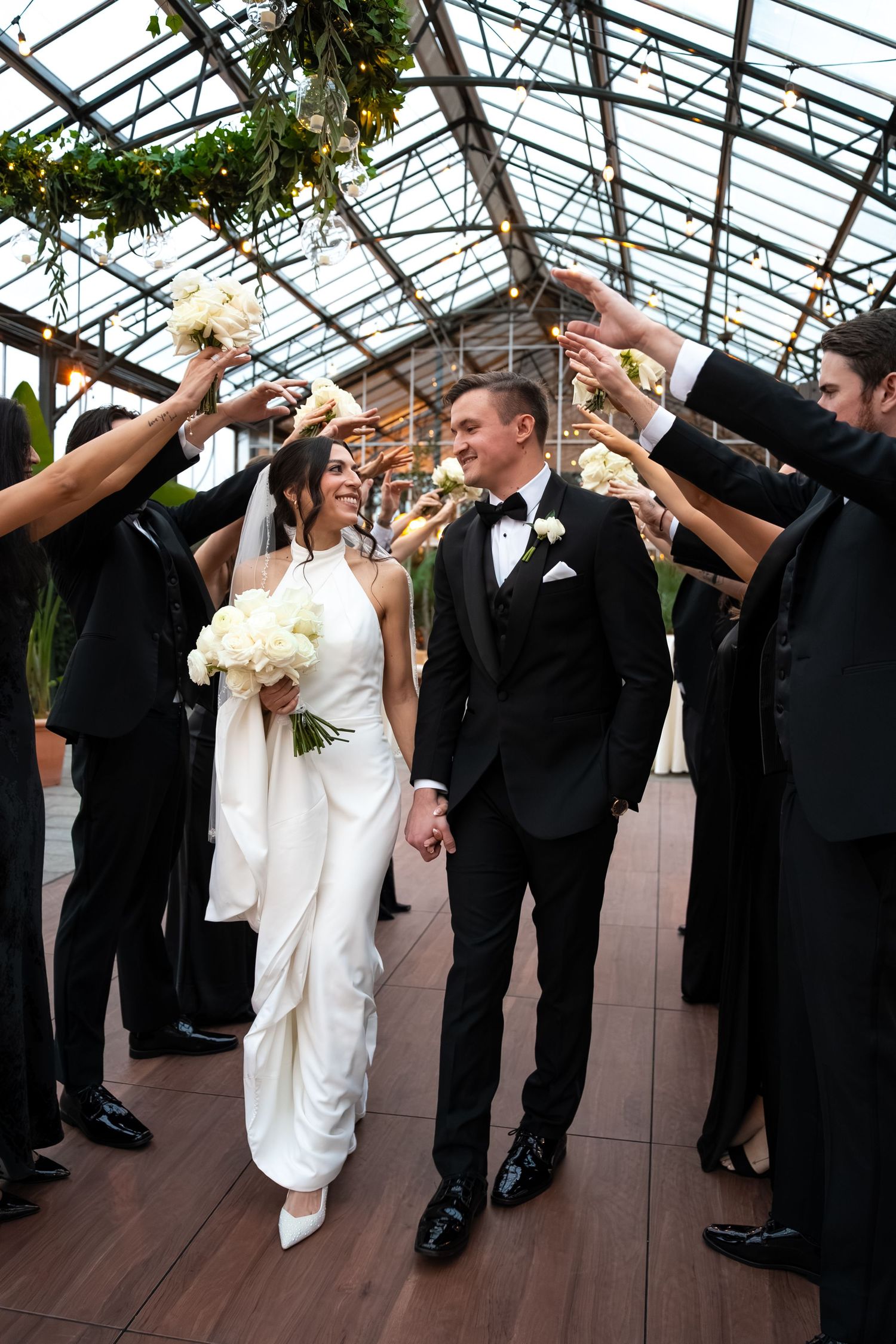 Bride and groom walk through floral arch tunnel held by wedding party in elegant greenhouse venue.