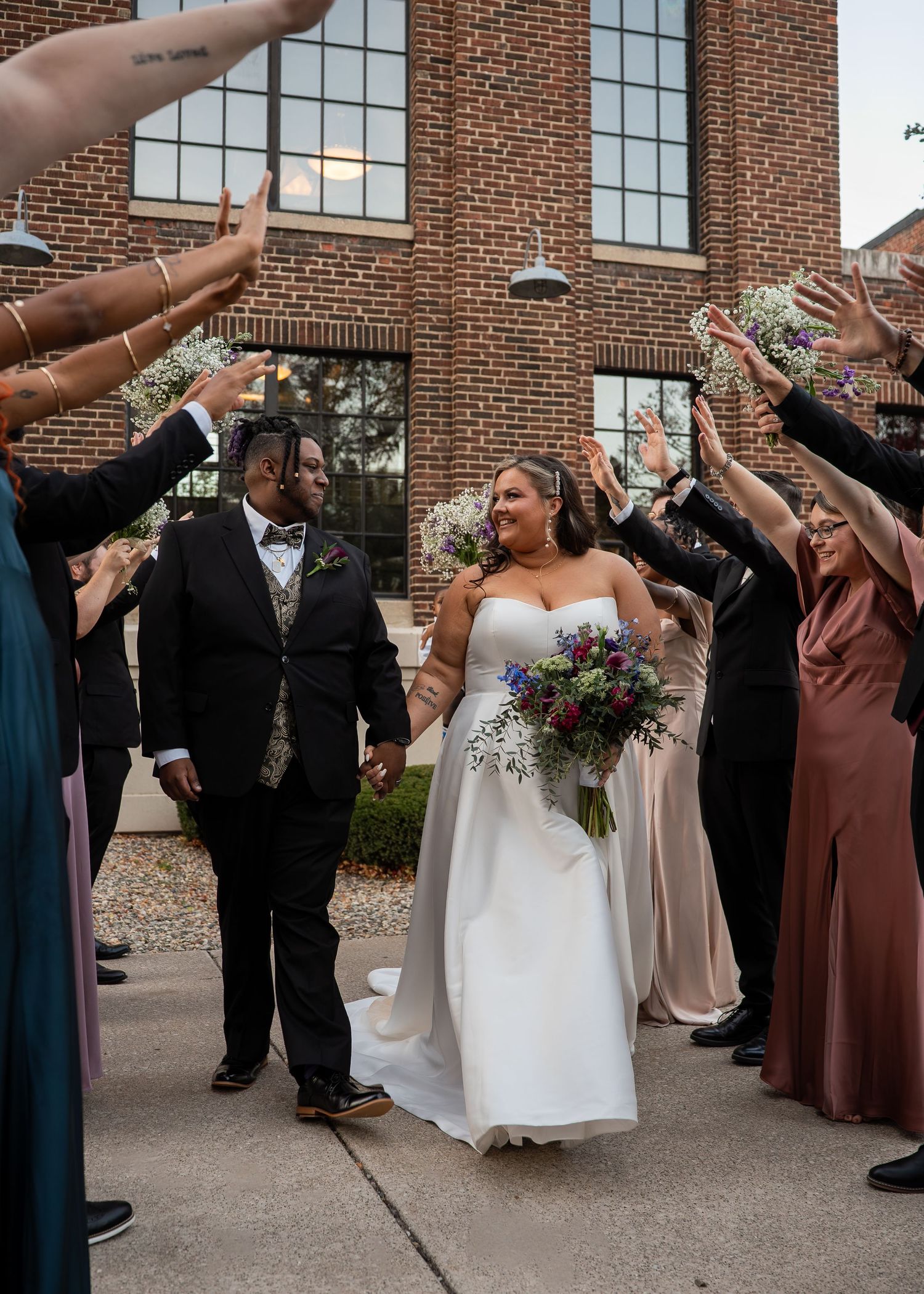 Bride and groom walk through a celebratory arch of raised hands outside a brick venue with wedding party.