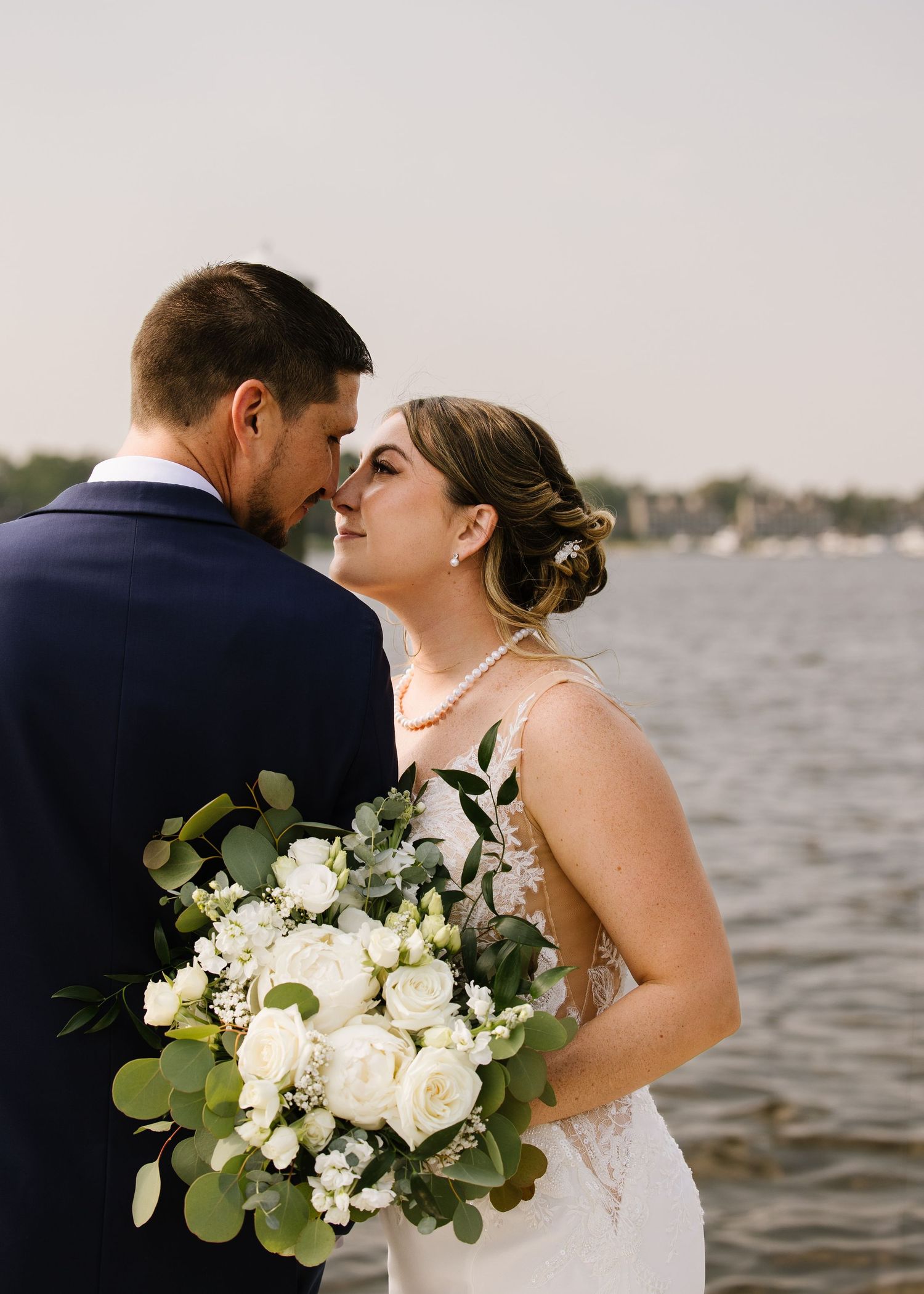 A couple shares an intimate moment by the water while holding a lush white and green wedding bouquet.