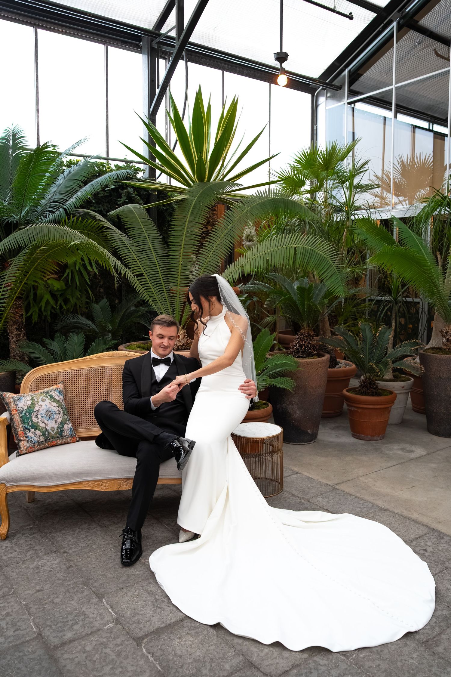Bride in white gown and groom in black tuxedo share a romantic moment on a vintage sofa in a lush tropical greenhouse.