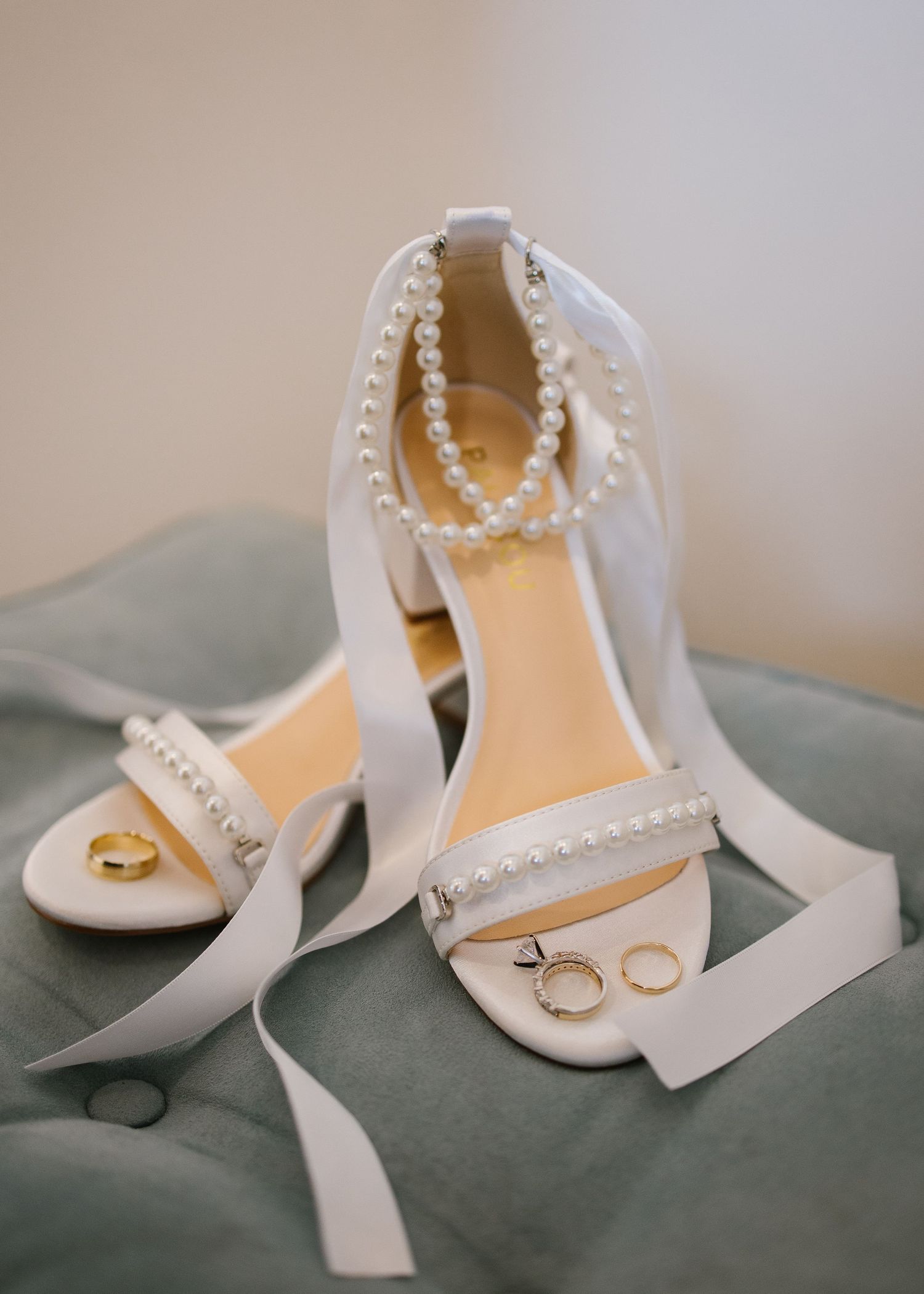 Elegant white bridal shoes with pearl straps and wedding rings displayed on sage green velvet fabric.